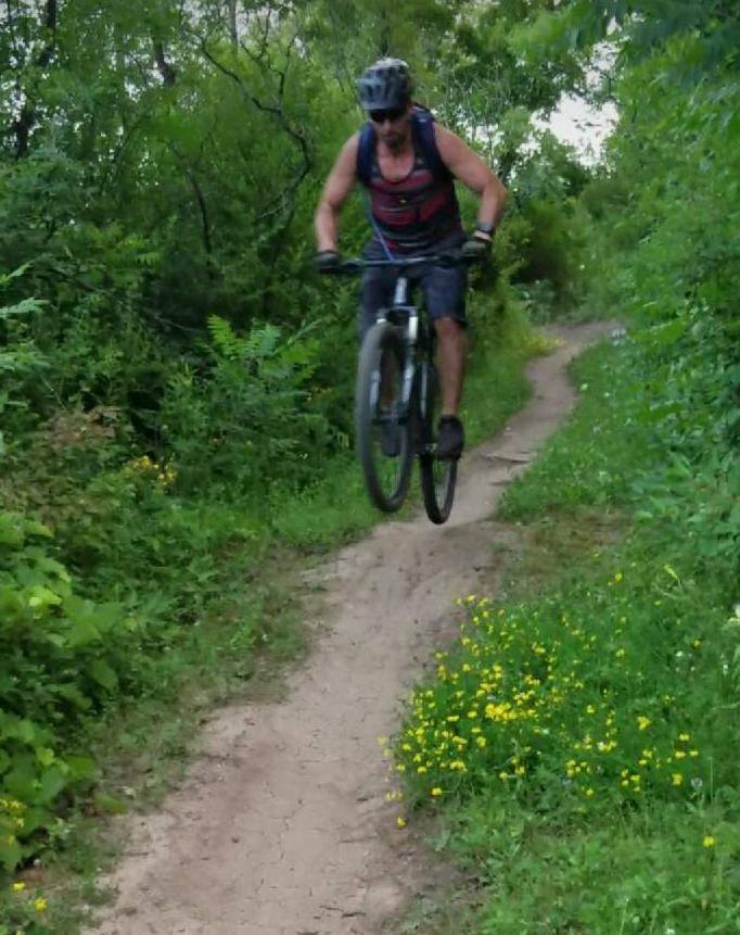 Specialized Rockhopper Comp Disc: A mountain biker in mid-air, jumping over a dirt trail surrounded by lush greenery and wildflowers. The cyclist is wearing a helmet and a sleeveless shirt, showcasing an action-packed moment in an outdoor setting.
