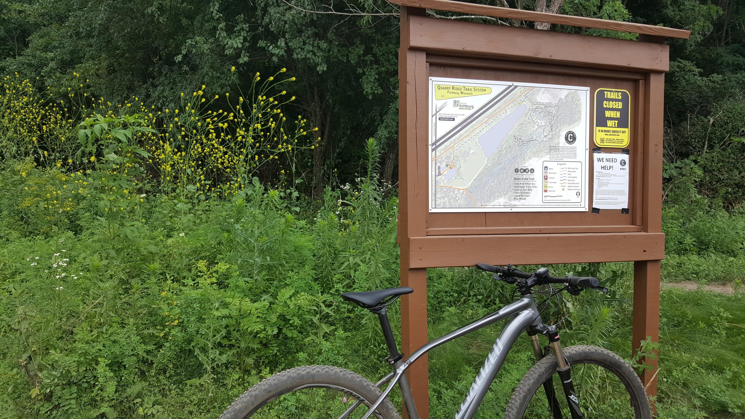 Specialized Rockhopper Comp Disc: A mountain bike leaning against a wooden trail sign displaying a map of the Quarry Ridge Trail System, surrounded by lush green vegetation and yellow wildflowers.
