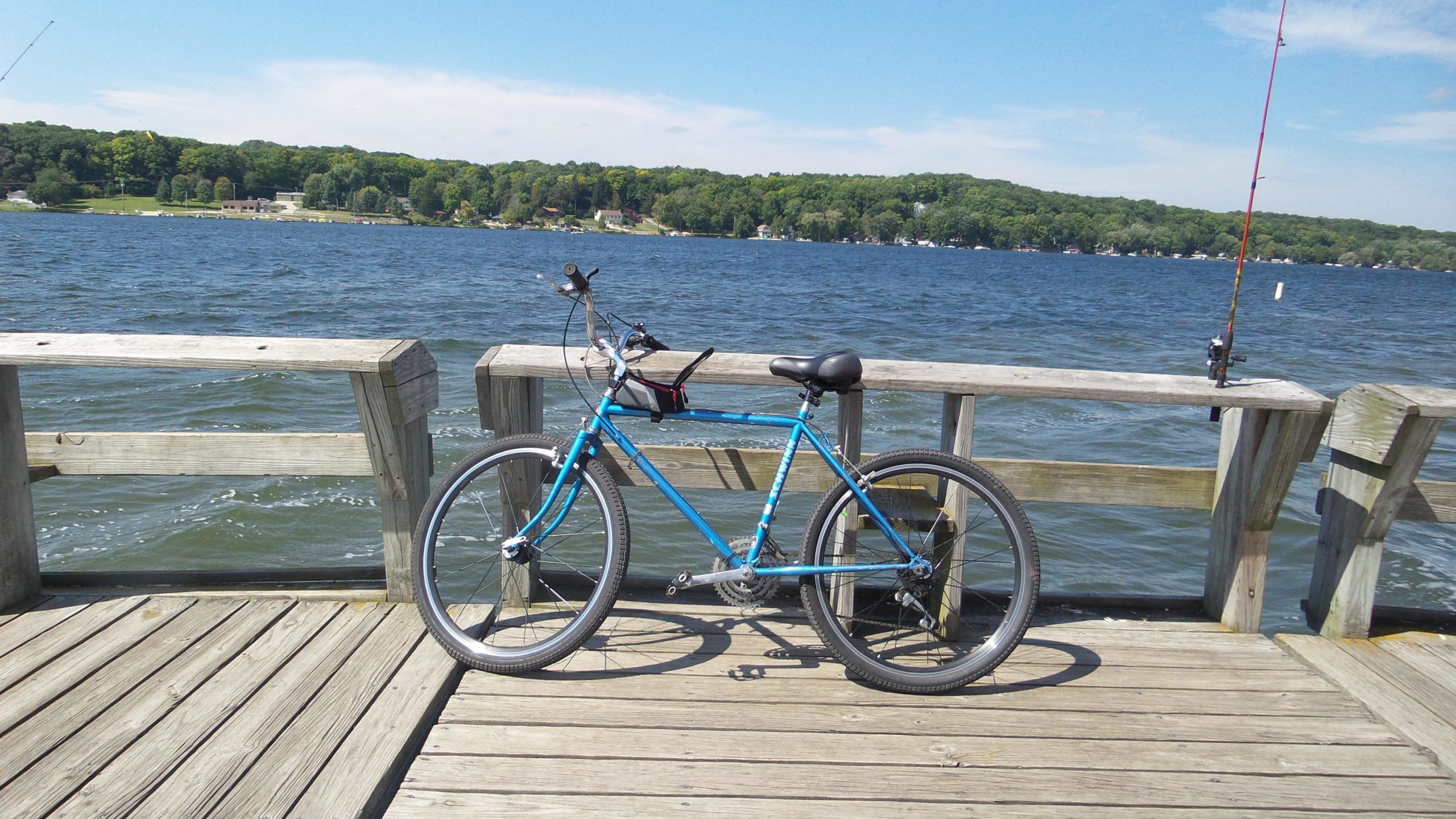 A blue bicycle parked on a wooden dock overlooking a lake, with a fishing rod propped against the railing and trees in the background under a clear blue sky. Lake to Lake mountain bike trail.