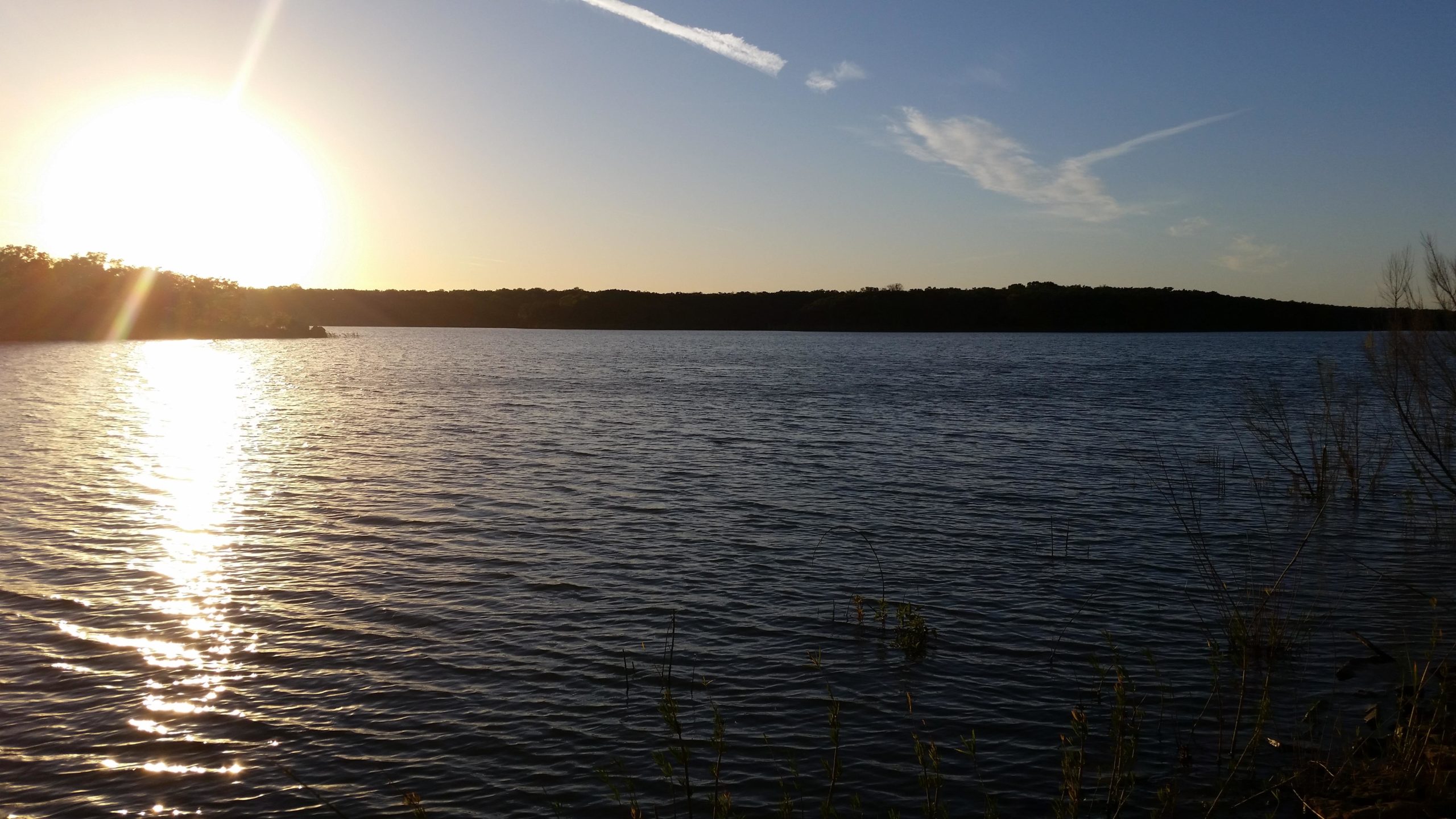 A serene lakeside scene during sunset, showcasing the sun setting low on the horizon, casting shimmering reflections on the water's surface. Gentle waves ripple across the lake, with trees visible in silhouette along the opposite shore under a clear blue sky. Lk Murray Section (park office to Tucker Tower) mountain bike trail.