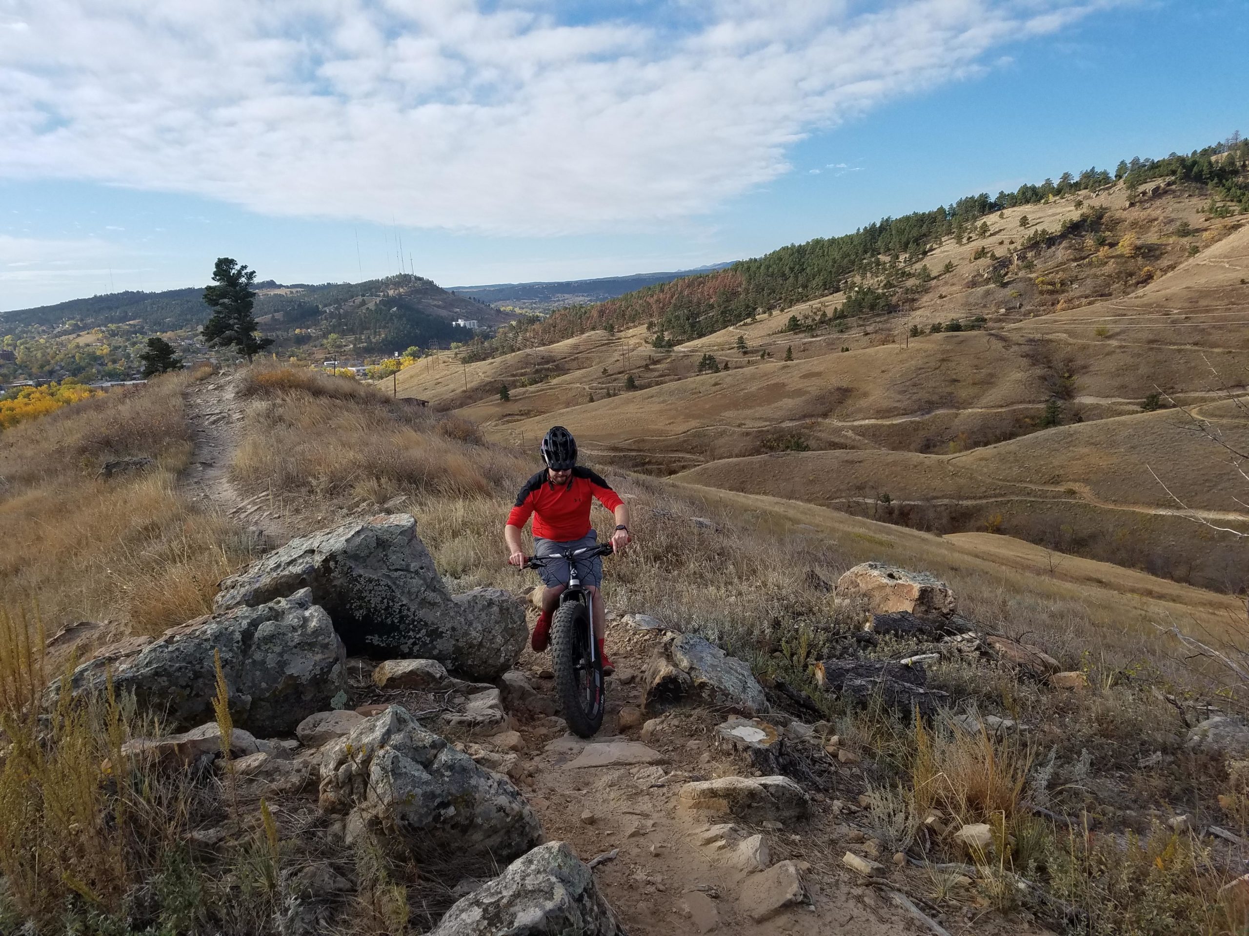 A person riding a fat tire bike on a rocky trail in a hilly landscape, surrounded by golden grass and autumn foliage. In the background, rolling hills and a clear blue sky create a scenic view. HLMP mountain bike trail.