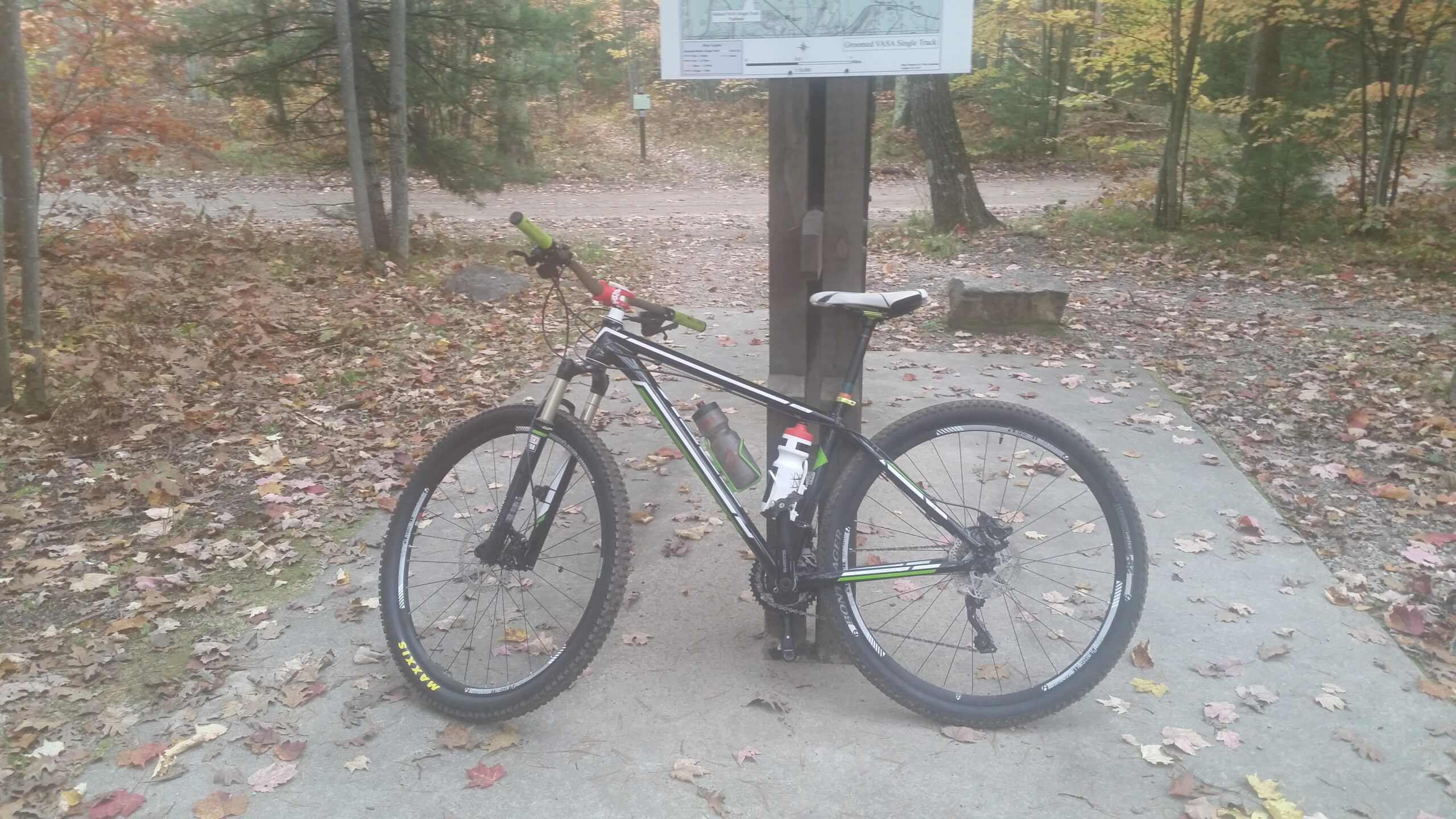 A black mountain bike with green accents and water bottles is parked on a concrete path surrounded by autumn leaves. In the background, there is a wooden signpost displaying a map. The scene is set in a wooded area with trees showcasing colorful fall foliage.