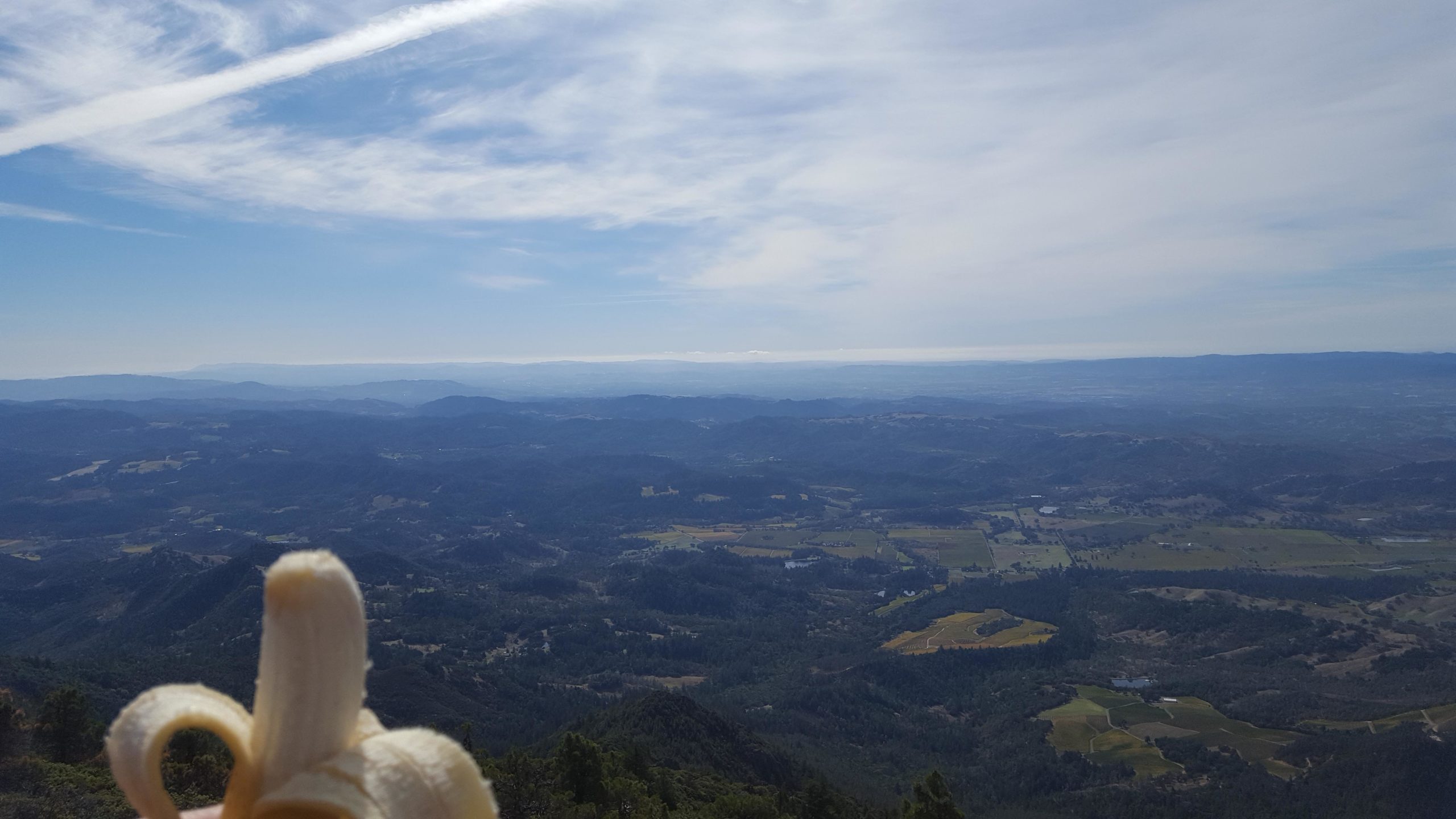 A banana being held in the foreground with a panoramic view of rolling hills and valleys under a partly cloudy sky in the background. Mount St Helena mountain bike trail.