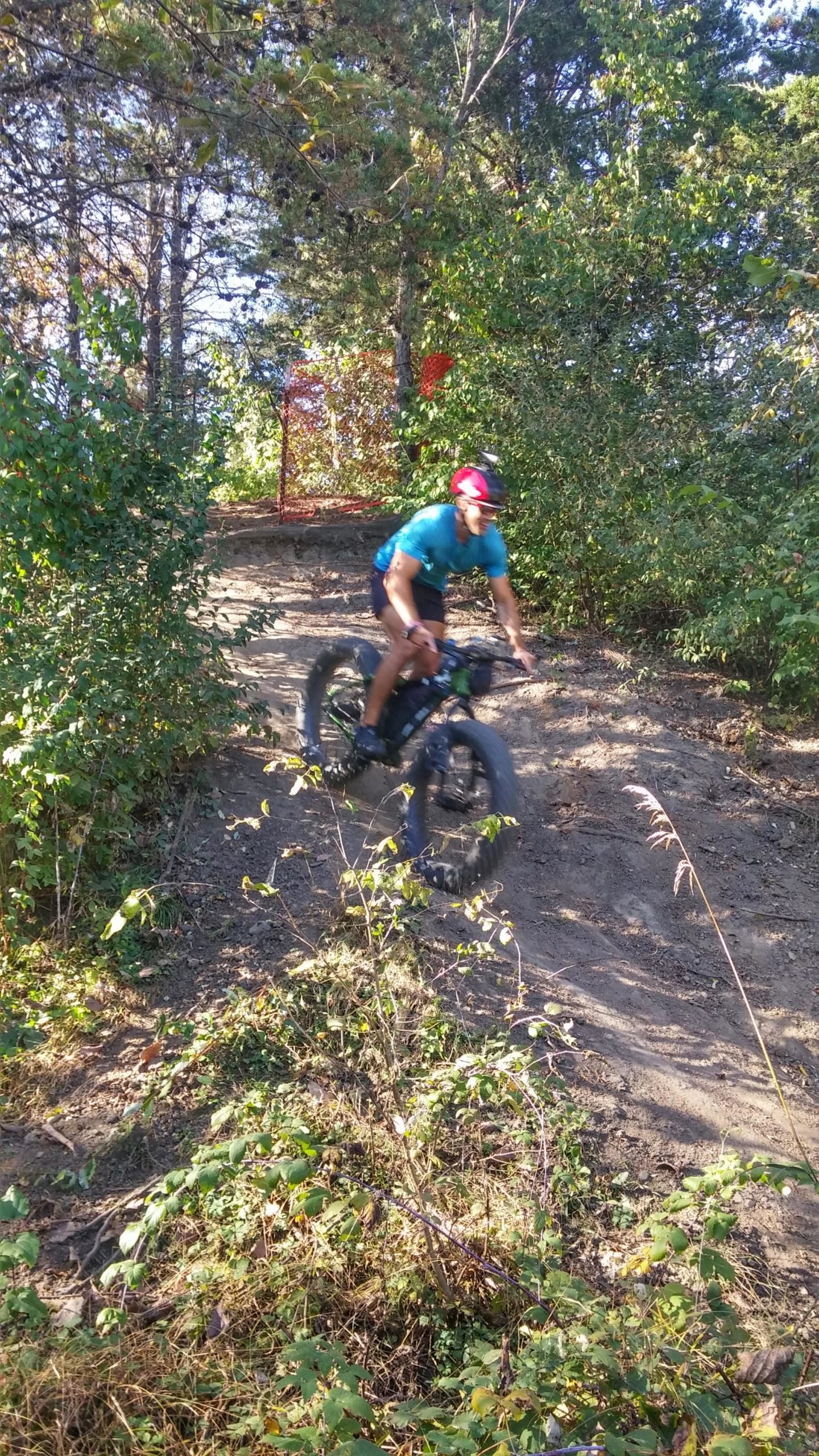 A cyclist riding a fat bike down a dirt trail in a wooded area, surrounded by greenery and sunlight filtering through the trees. The cyclist is wearing a blue shirt and a helmet, with some vegetation in the foreground. Banner Lakes At Summerset Park mountain bike trail.