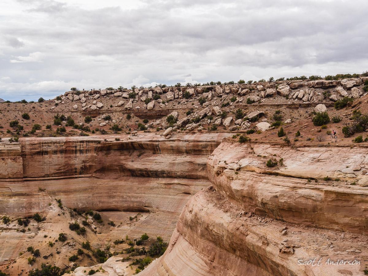 A panoramic view of a rugged canyon landscape featuring layered rock formations and a cloudy sky. The foreground showcases smooth, eroded cliffs with patches of greenery, while the background includes a rocky ridge lined with shrubs. A small figure can be seen near the edge of the canyon, adding a sense of scale to the vast natural scenery. Western Rim mountain bike trail.