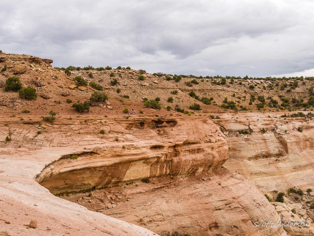 A rugged, rocky landscape with layered hills and sparse vegetation under a cloudy sky. A small figure can be seen biking along a cliff edge, showcasing the vastness and natural beauty of the terrain. Western Rim mountain bike trail.