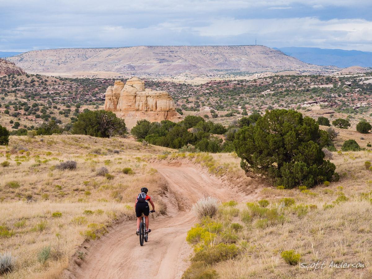 A mountain biker riding along a dirt trail in a rugged, desert landscape, with prominent rock formations and rolling hills in the background under a partly cloudy sky. Rabbit Valley mountain bike trail.