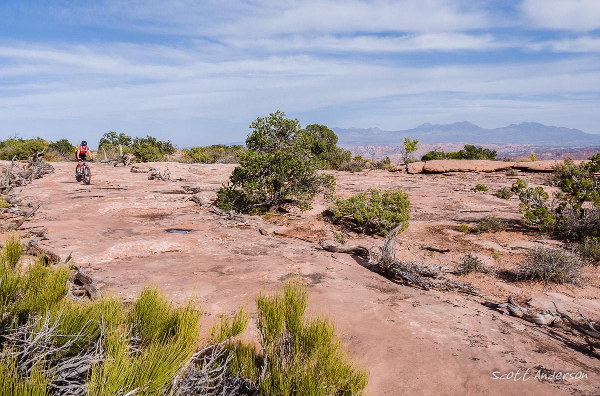 A mountain biker rides along a rocky trail surrounded by sparse vegetation and low bushes under a partly cloudy sky. In the background, distant mountains are visible, creating a scenic outdoor landscape. Dead Horse Point State Park mountain bike trail.
