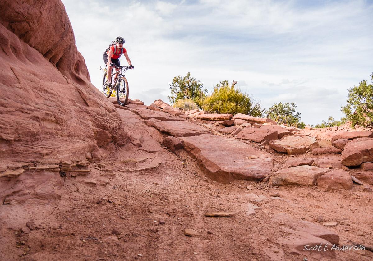 Mountain biker navigating a rocky terrain on a red rock landscape under a partly cloudy sky. The rider is focused, wearing a helmet and cycling gear, as they maneuver over uneven ground surrounded by low vegetation. Dead Horse Point State Park mountain bike trail.