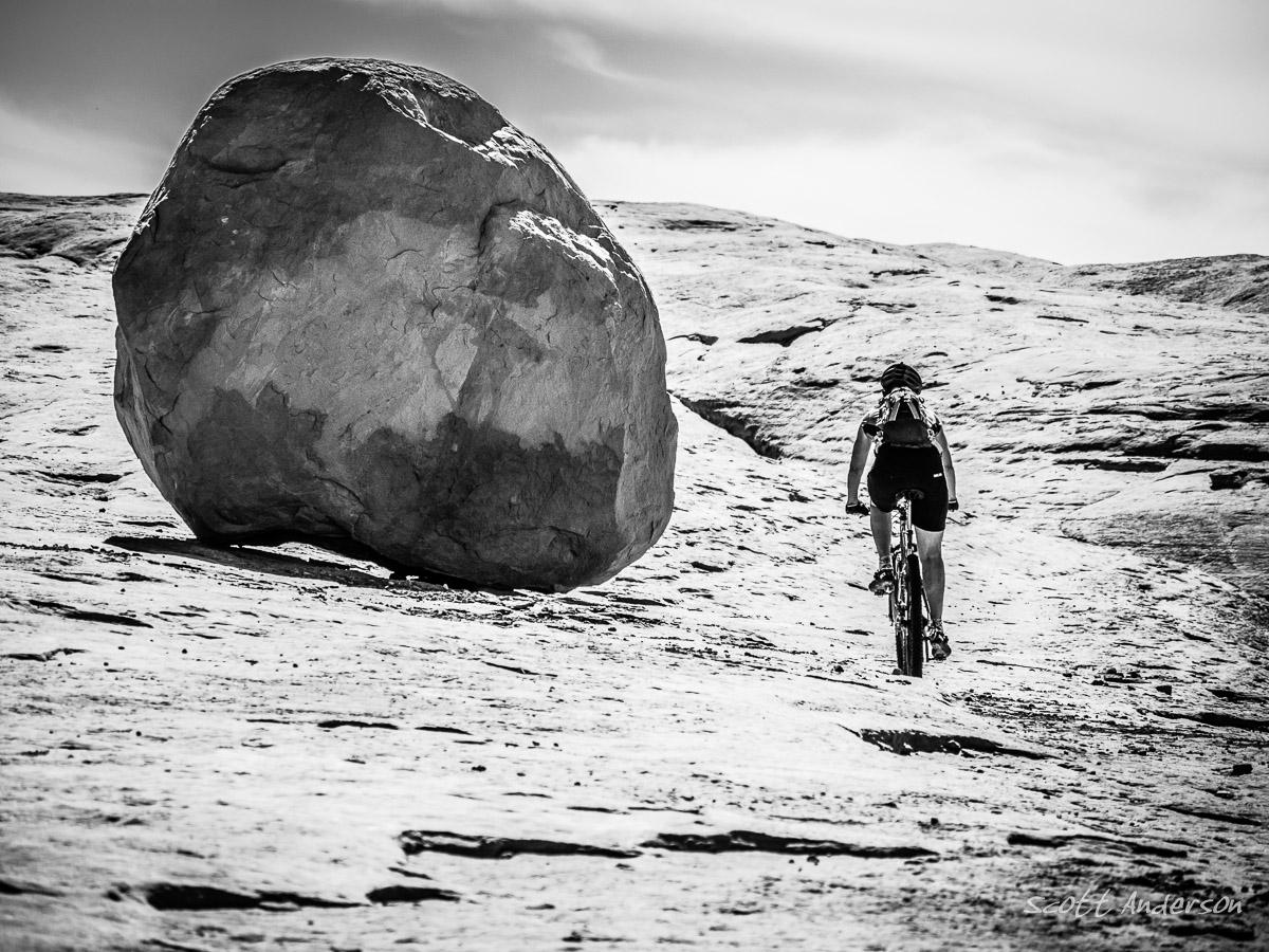 A mountain biker riding on a rocky, textured landscape, with a large boulder to the side. The image is presented in black and white, emphasizing the natural features of the terrain and the cyclist's silhouette against the sky. Monitor & Merrimac Singletrack mountain bike trail.