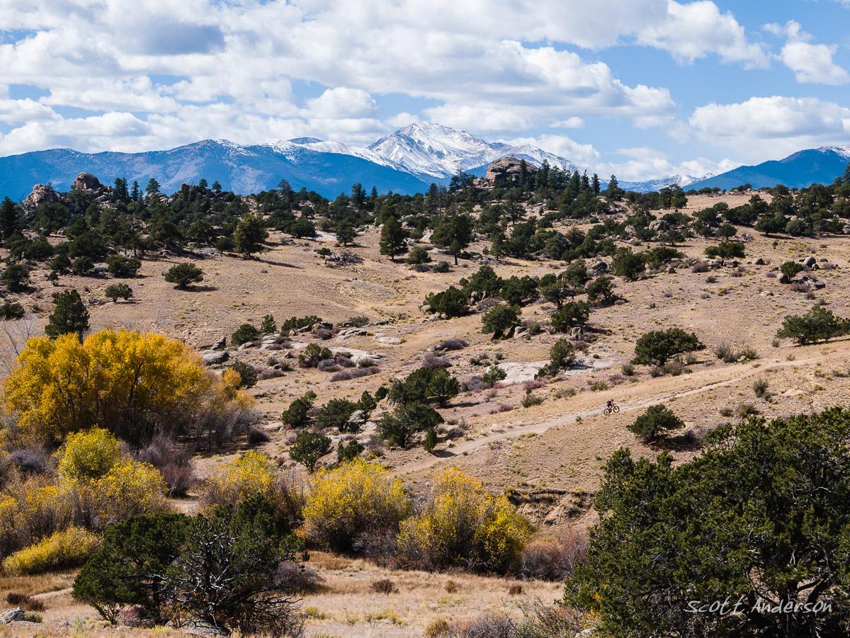 A scenic view of a mountainous landscape featuring green trees and golden autumn foliage in the foreground, leading up to snow-capped peaks under a partly cloudy blue sky. A dirt path winds through the terrain, where a cyclist can be seen riding in the distance. Trail #1414 mountain bike trail.