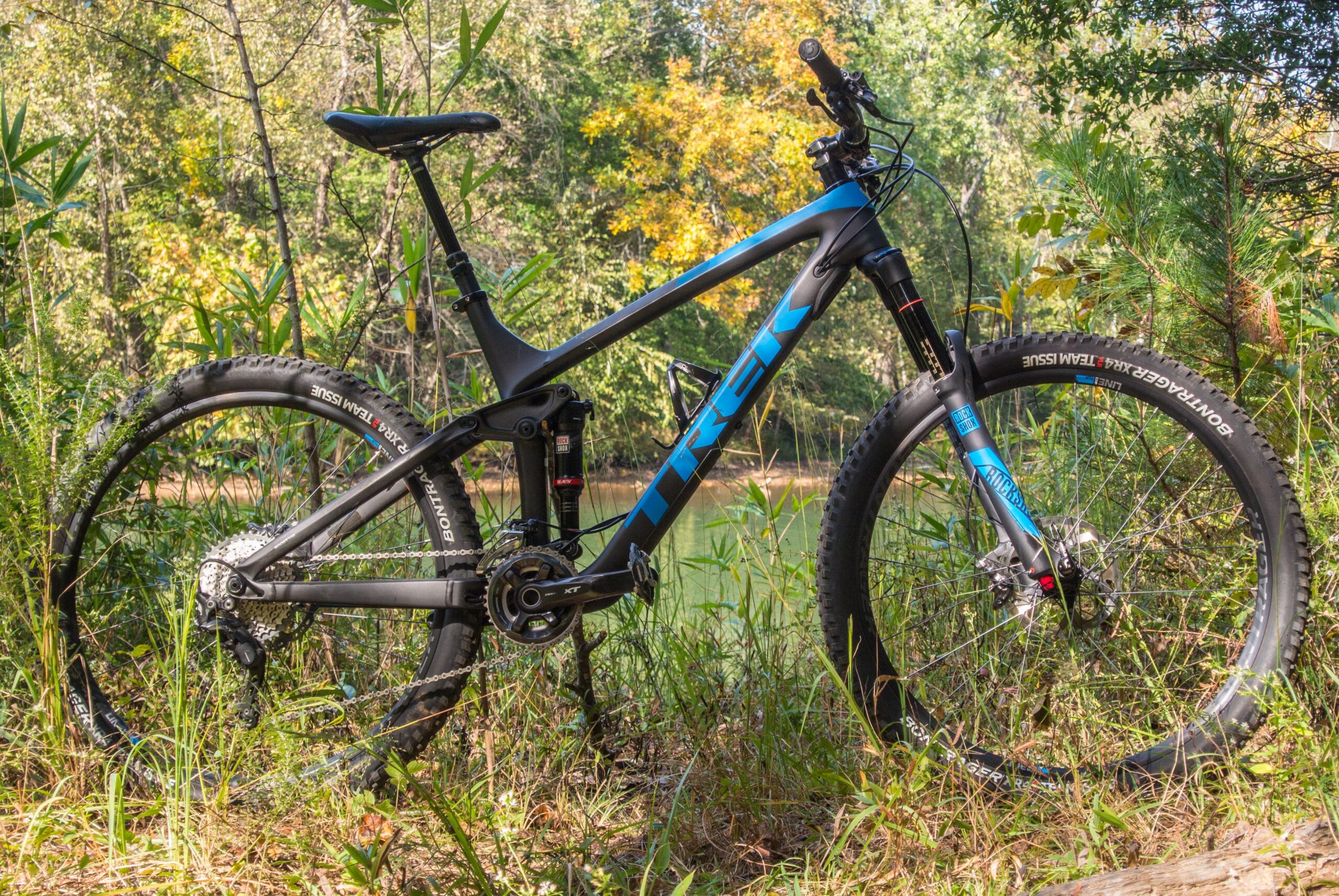 Trek Remedy 9: A black and blue Trek mountain bike stands in a natural setting with greenery and a glimpse of water in the background. The bike is positioned amongst tall grass and plants, showcasing its design and features.