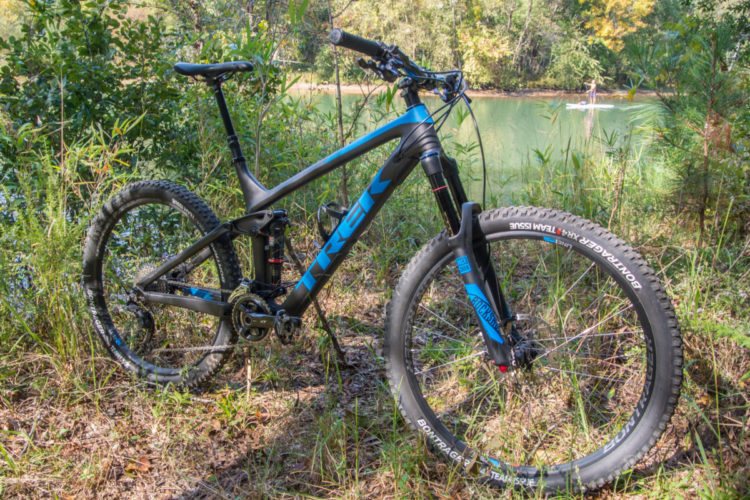 Mountain bike positioned on grass near a tranquil lake, surrounded by lush greenery. The bike features a sleek black frame with blue accents, highlighting its brand name. Sunlight filters through the trees, creating a picturesque outdoor setting perfect for cycling adventures.
