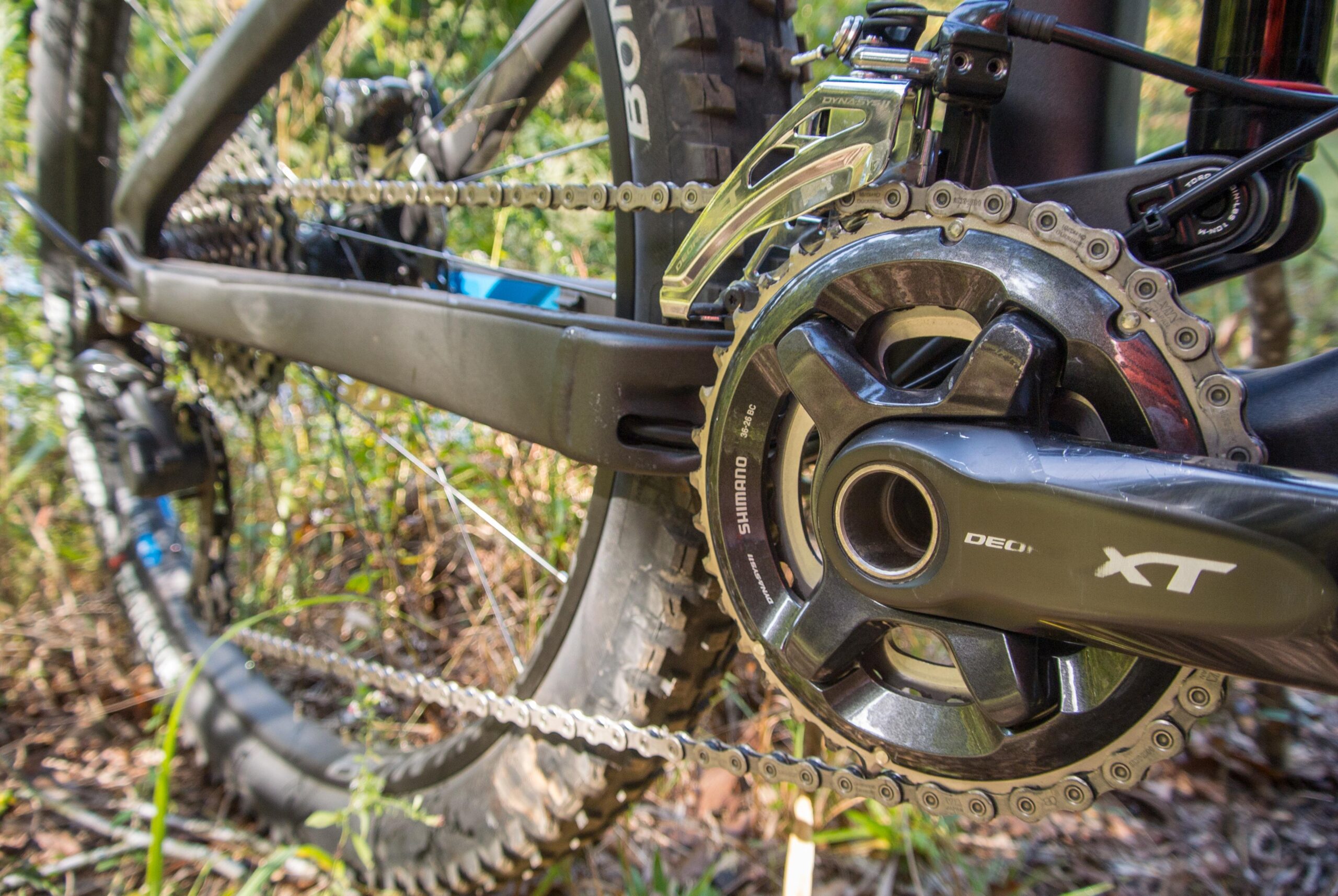 Trek Remedy 9: Close-up image of a mountain bike's drivetrain, featuring a Shimano XT crankset and chain, set against a natural background with foliage. The focus highlights the gears, chain, and bike frame details.