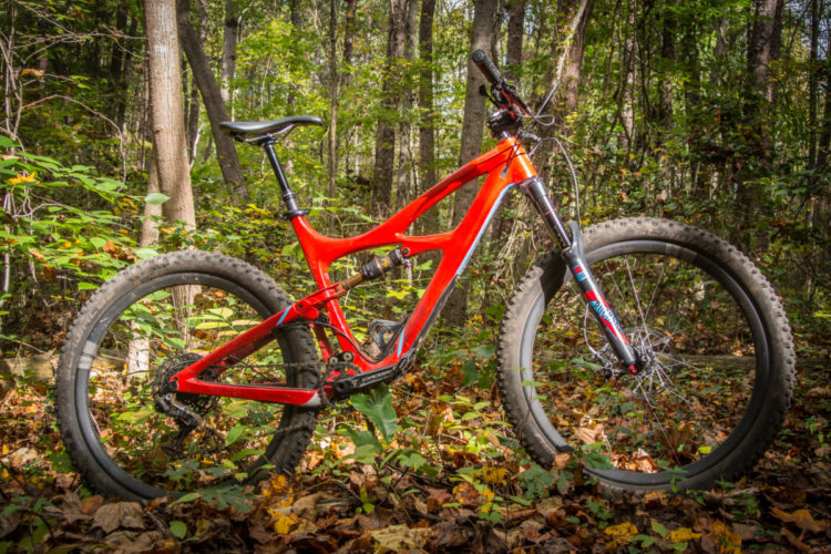 A vibrant red mountain bike positioned among fallen leaves and greenery in a forest setting, showcasing its rugged design and thick tires. Sunlight filters through the trees, illuminating the bike's frame and components.