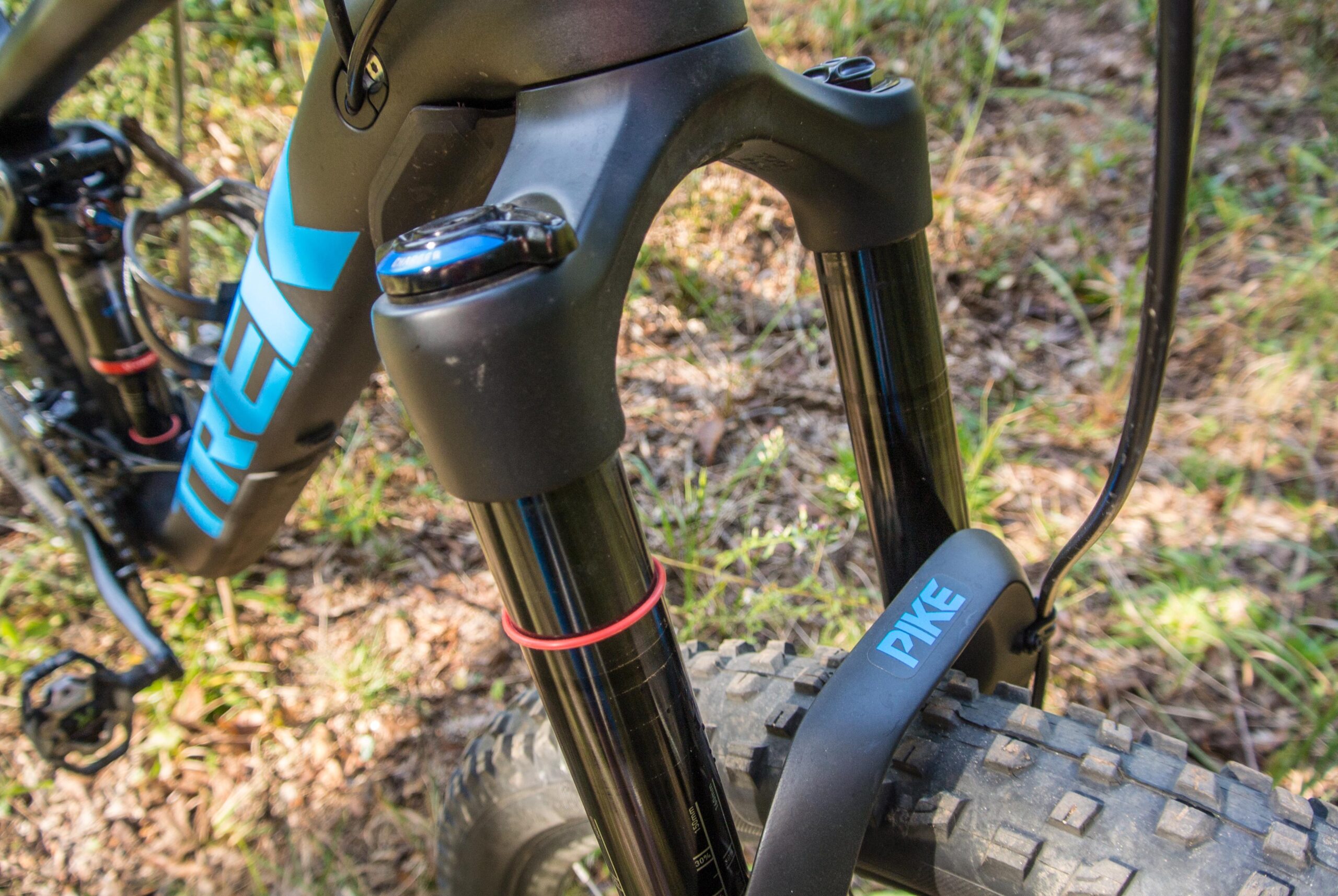 Trek Remedy 9: Close-up view of a mountain bike's front fork, featuring the Trek logo prominently displayed on the frame and the Pike branding on the fork. The background includes natural foliage and ground cover, suggesting an outdoor setting.