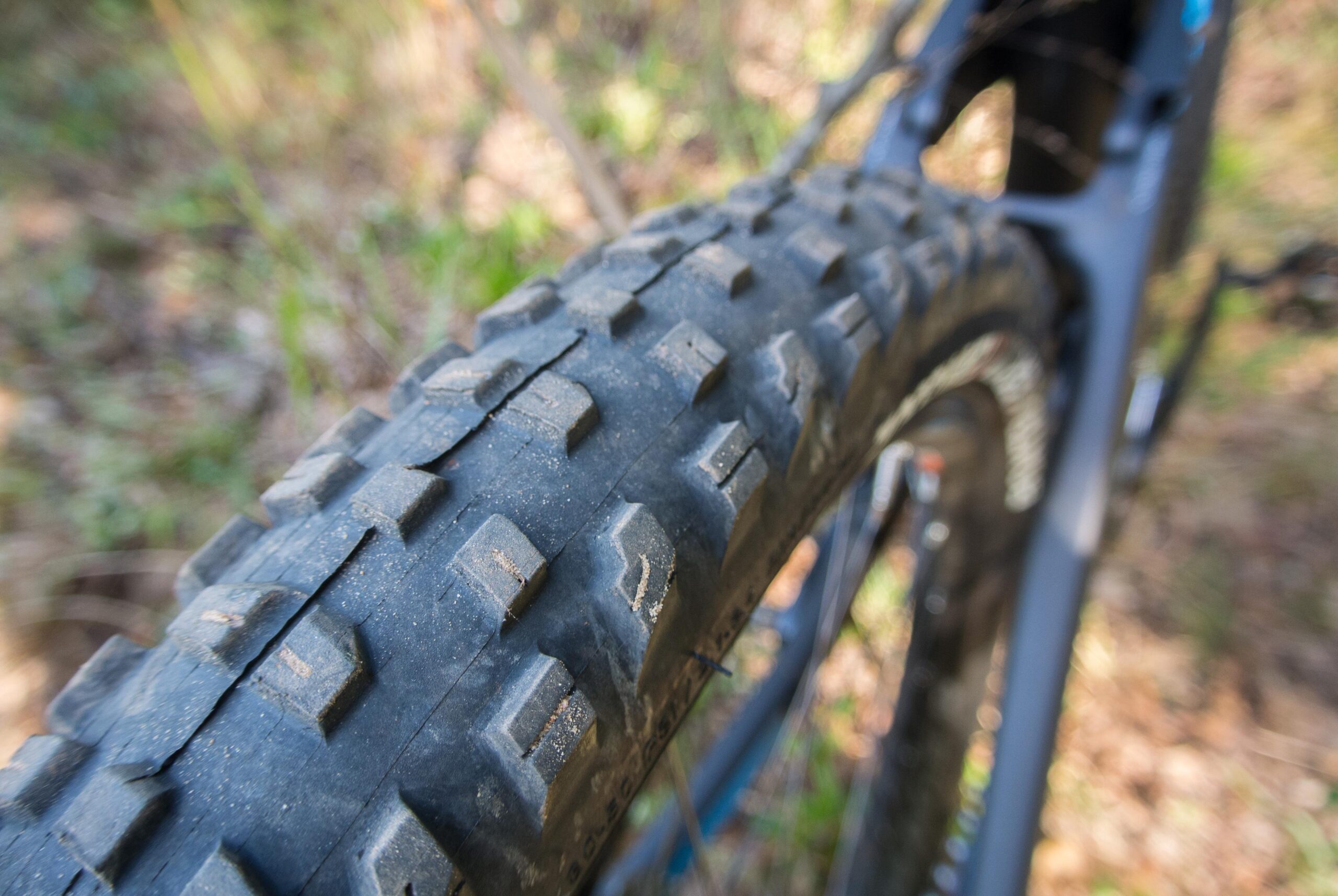 Trek Remedy 9: Close-up view of a mountain bike tire showcasing its tread pattern, with a blurred background of greenery and natural terrain. The tire appears slightly worn, indicating use on rugged trails.
