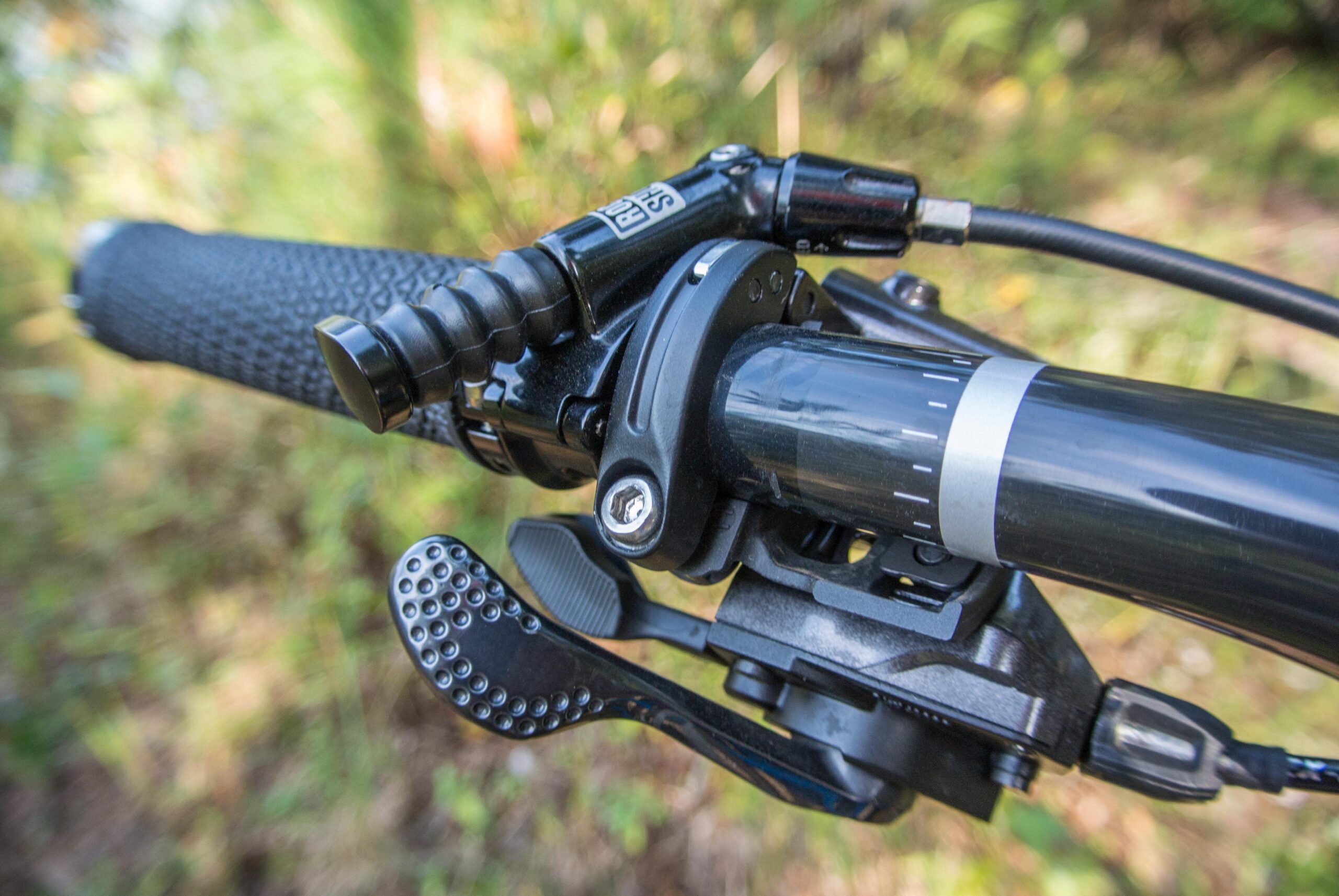 Trek Remedy 9: Close-up view of a mountain bike handlebar, featuring textured grips, brake and shifter controls, and a lever mechanism, set against a blurred natural background.