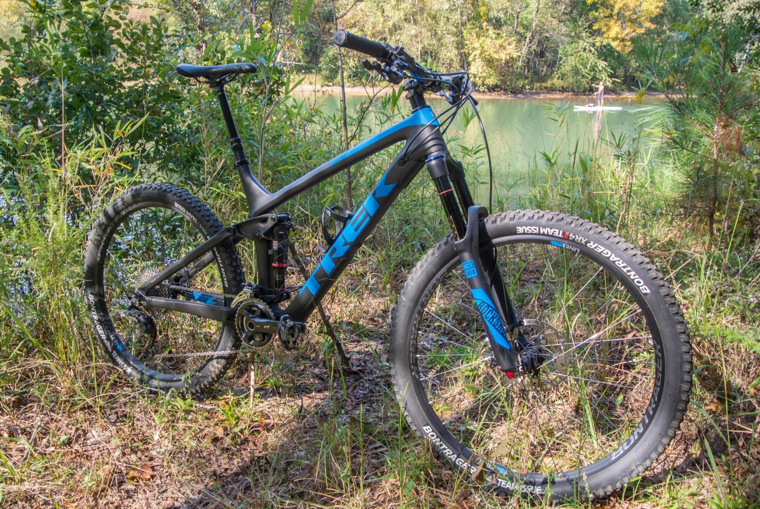 Trek Remedy 9: A black and blue mountain bike parked on the edge of a wooded area, with green foliage in the background and a calm body of water visible in the distance. The bike features a sleek frame, wide tires, and a suspension system, suggesting it is designed for off-road trails.