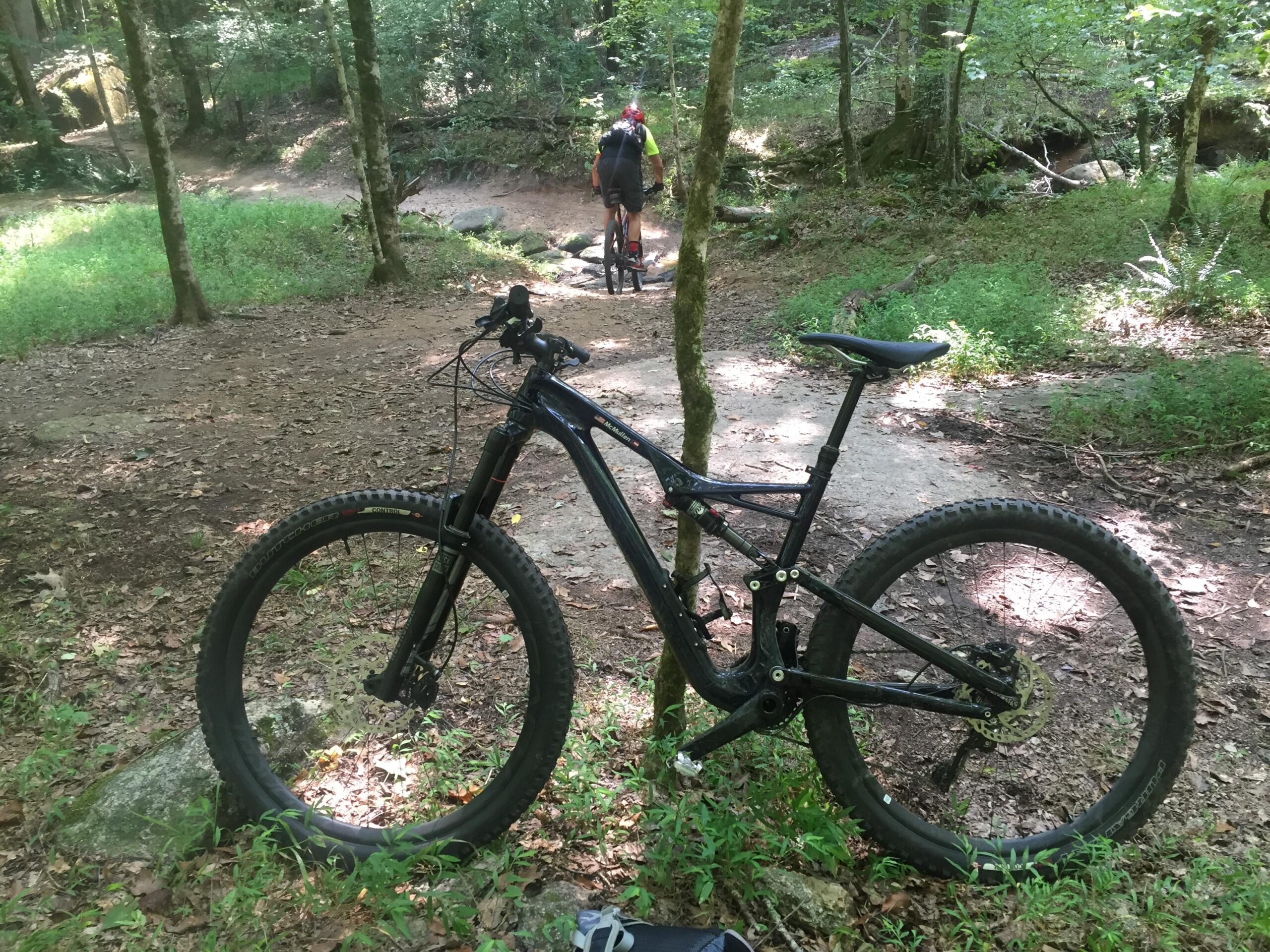Specialized Stumpjumper FSR comp carbon 650B: A close-up view of a mountain bike resting on the ground in a wooded area, with a blurred cyclist riding along a dirt path in the background. The scene features lush green foliage and sunlight filtering through the trees.
