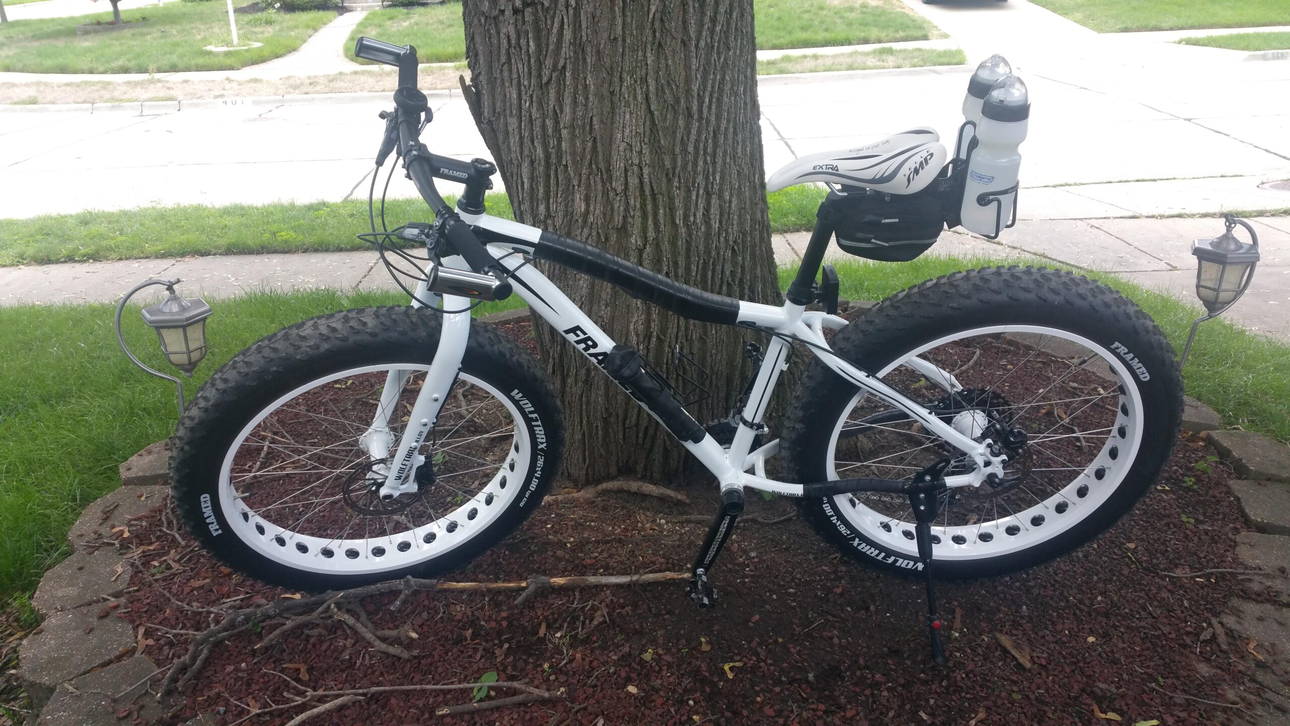 Framed Framed Wolftrax: A white and black fat tire bike leaning against a tree, with a water bottle holder mounted on the back. The bike features wide tires with white rims and has a stand deployed. In the background, there's a grassy area and a lamp post near a sidewalk.