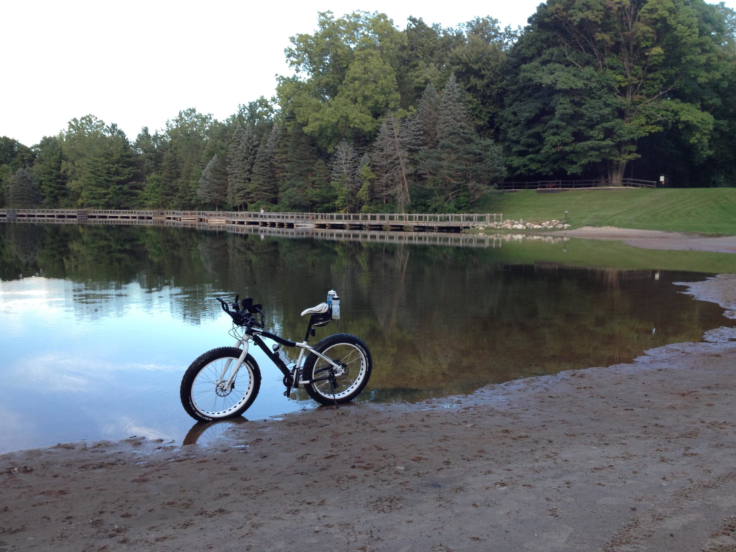 A bicycle resting on the sandy shore of a calm lake, with reflections of trees in the water. In the background, a wooden boardwalk runs alongside the lake, bordered by lush green foliage and a grassy area. The scene captures a peaceful outdoor setting. Burchfield mountain bike trail.