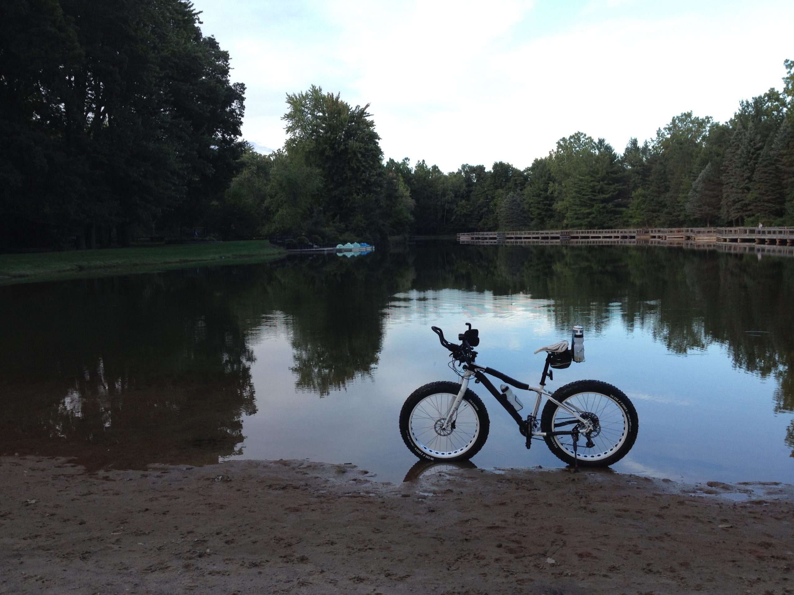A mountain bike is parked on a sandy shore near a calm lake, surrounded by lush green trees. The water reflects the scenery, creating a serene and tranquil atmosphere. In the background, a wooden walkway can be seen along the edge of the lake, with a few paddle boats moored nearby. The overall mood is peaceful and inviting, ideal for outdoor activities. Burchfield mountain bike trail.