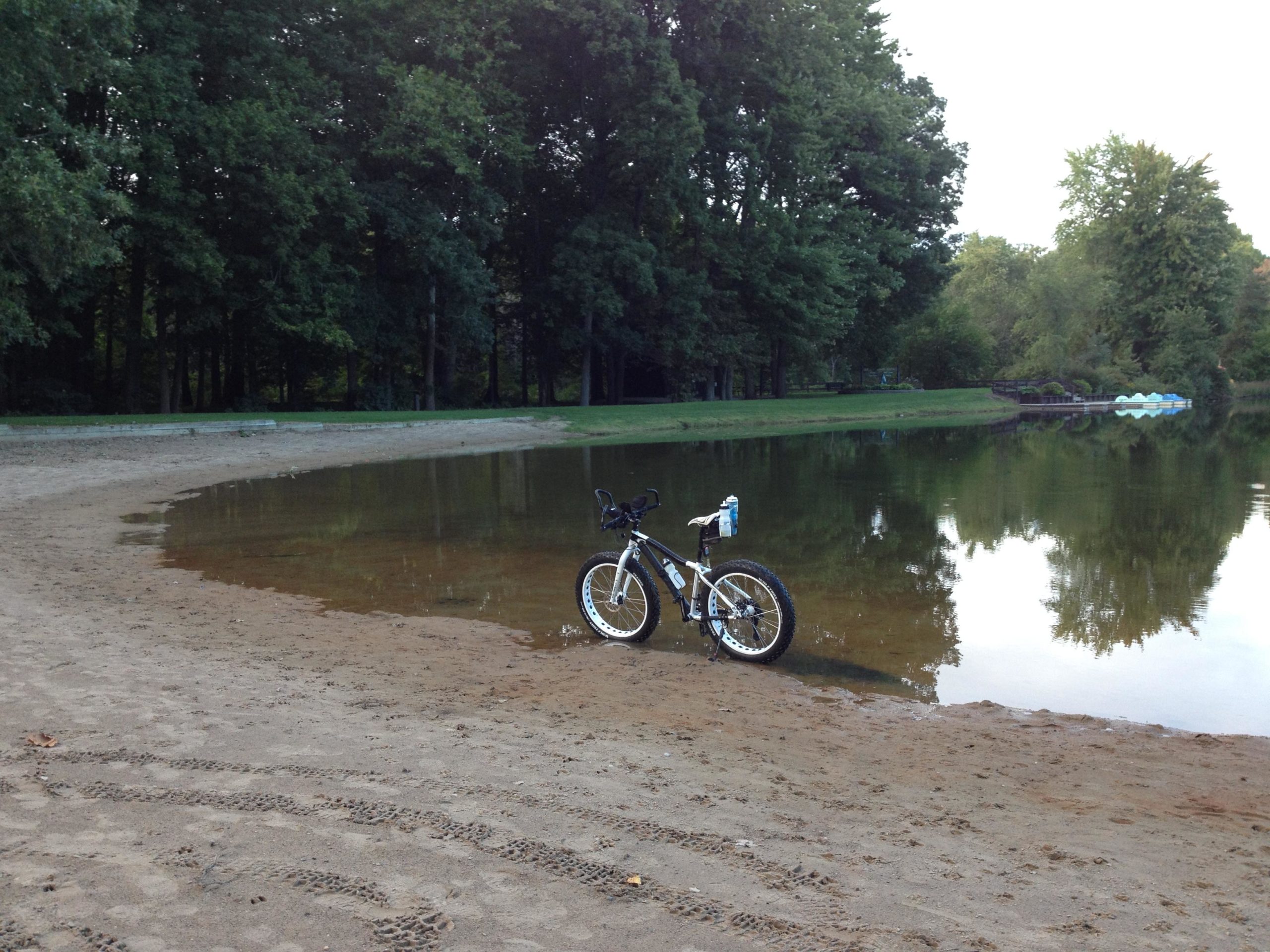 A mountain bike is parked on a sandy shoreline by a calm lake, surrounded by lush green trees. The water reflects the trees, creating a tranquil atmosphere. In the background, a small dock with paddle boats can be seen. Burchfield mountain bike trail.