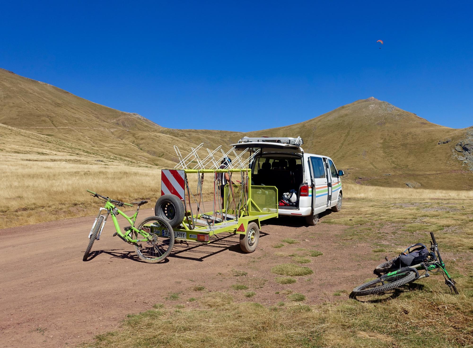 A van parked on a dirt road in a mountainous area, with a green bicycle and a bicycle trailer loaded with equipment. The landscape features grassy hills under a clear blue sky. A paraglider can be seen flying in the distance. Gallinero mountain bike trail.