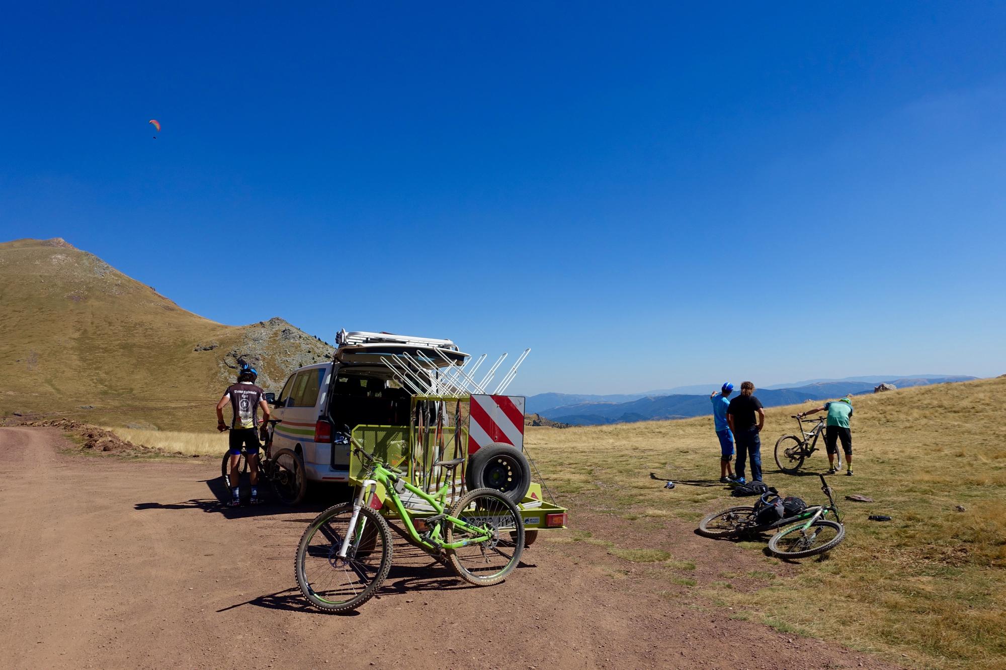A group of three people stands near a parked van on a dirt road in a mountainous area. Two individuals are working on their bicycles, while one person observes. A vibrant, orange paraglider is visible in the clear blue sky above. Nearby, several mountain bikes are resting on the ground, surrounded by dry grass and hills. Gallinero mountain bike trail.