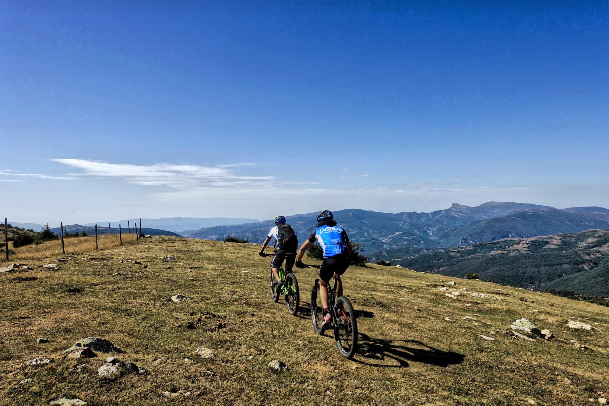 Two cyclists riding along a grassy path with a mountainous landscape in the background under a clear blue sky. The terrain is rocky and hilly, and the scene conveys a sense of adventure and outdoor activity. Serra Espina mountain bike trail.