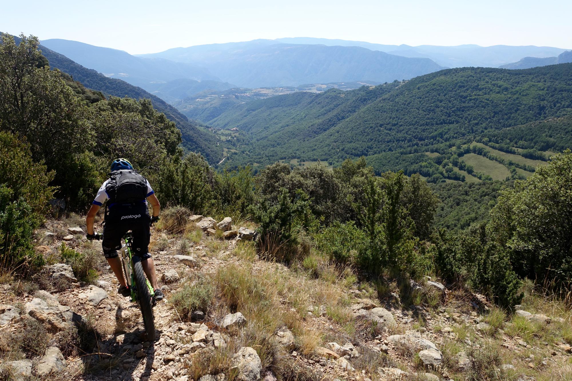 A mountain biker navigating a rocky trail surrounded by lush green hills and a expansive valley in the background under a clear blue sky. Serra Espina mountain bike trail.
