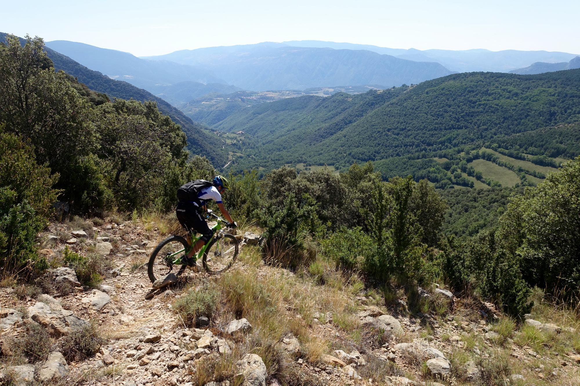 A mountain biker riding down a rocky trail with a scenic view of green hills and valleys in the background under a clear blue sky. Serra Espina mountain bike trail.
