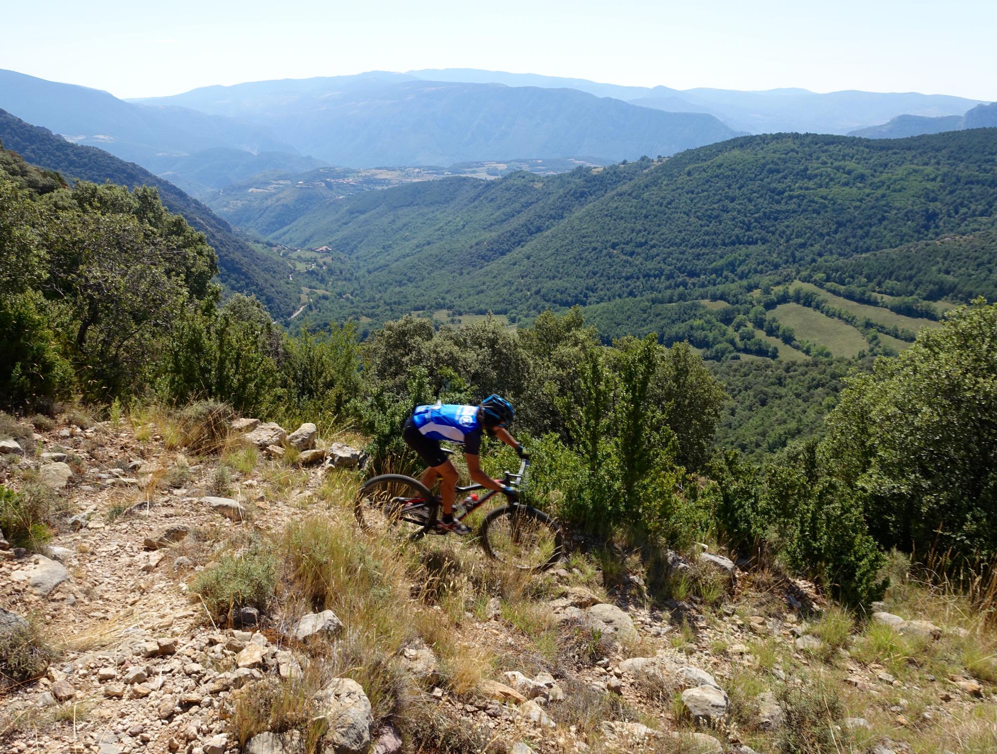 A mountain biker navigates a rocky trail along a hillside, surrounded by lush green vegetation and distant mountains under a clear blue sky. The scene captures the thrill of outdoor adventure in a scenic landscape. Serra Espina mountain bike trail.