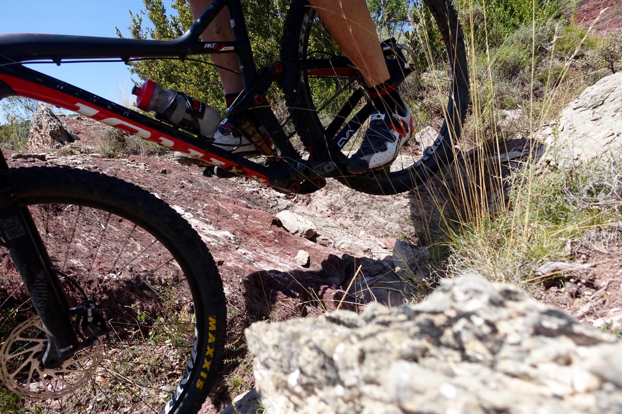 A close-up view of a mountain bike navigating a rocky terrain. The bike's frame is prominently displayed with a water bottle mounted on it. One foot is positioned on the pedal while the other hovers above a rough, uneven surface made up of large rocks and grass. The background features greenery and blue sky, suggesting an outdoor adventure setting. Serra Espina mountain bike trail.