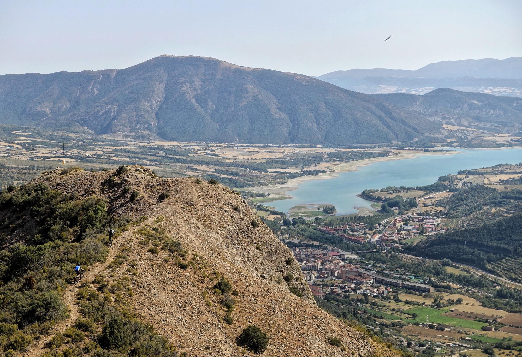 A panoramic view of a mountainous landscape featuring a winding trail along a rocky ridge. In the foreground, two cyclists navigate the trail, while the background showcases rolling hills, a serene lake, and a small town nestled in the valley below, surrounded by farmland and greenery under a clear blue sky. Cresta De Gelat mountain bike trail.