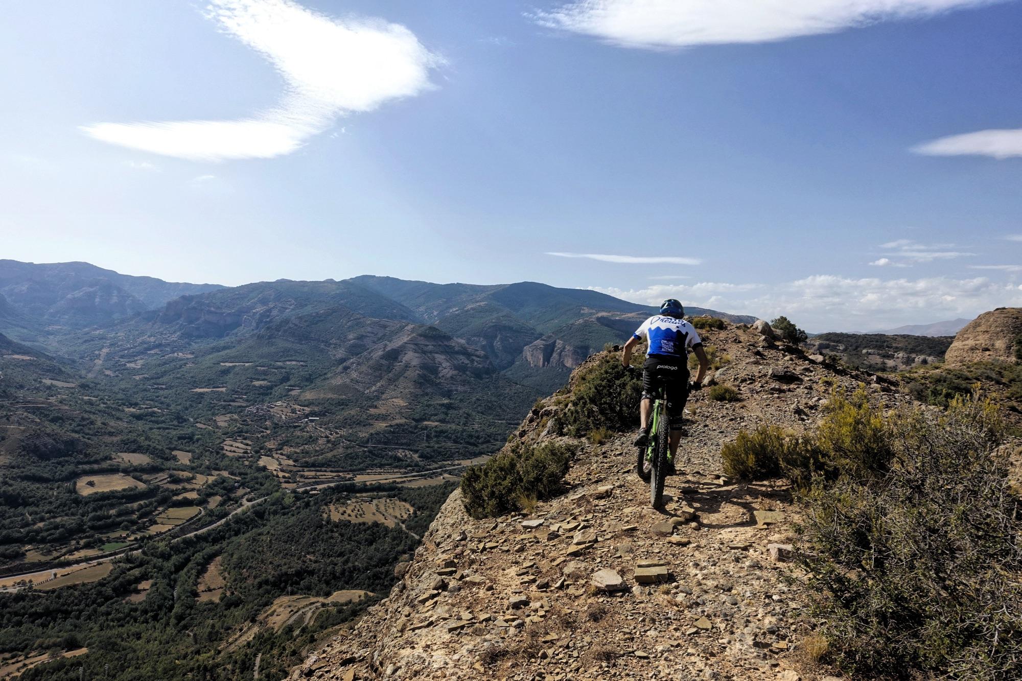 A mountain biker riding along a rocky trail on a hillside, with expansive green valleys and mountains in the background under a clear blue sky. Cresta De Gelat mountain bike trail.