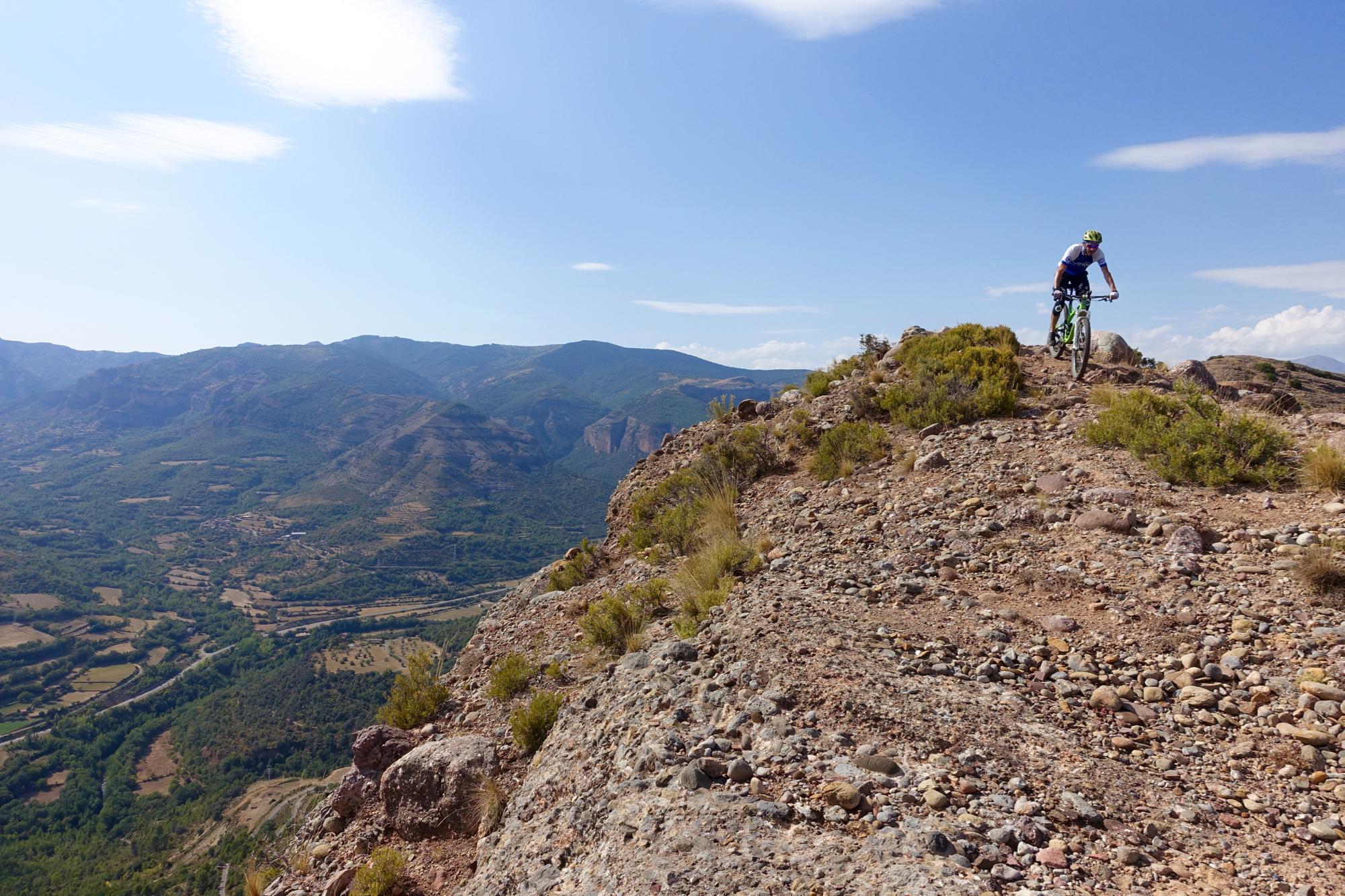 A mountain biker navigating a rocky trail on a mountainside, with expansive green valleys and distant mountains visible in the background under a clear blue sky. Cresta De Gelat mountain bike trail.