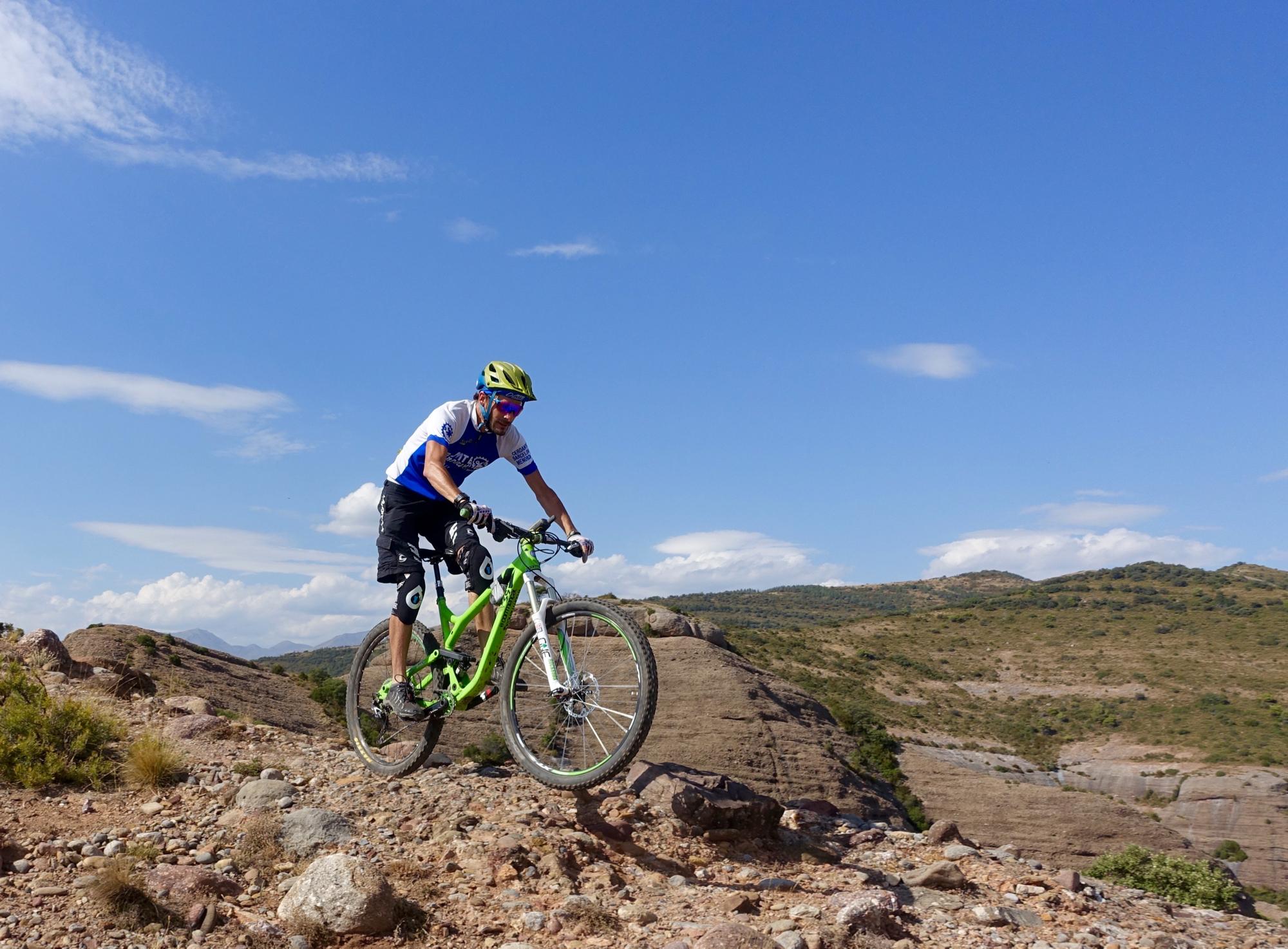 A mountain biker in a blue and white jersey, wearing a helmet and sunglasses, jumps off a rocky terrain, with hills and a clear blue sky in the background. Cresta De Gelat mountain bike trail.