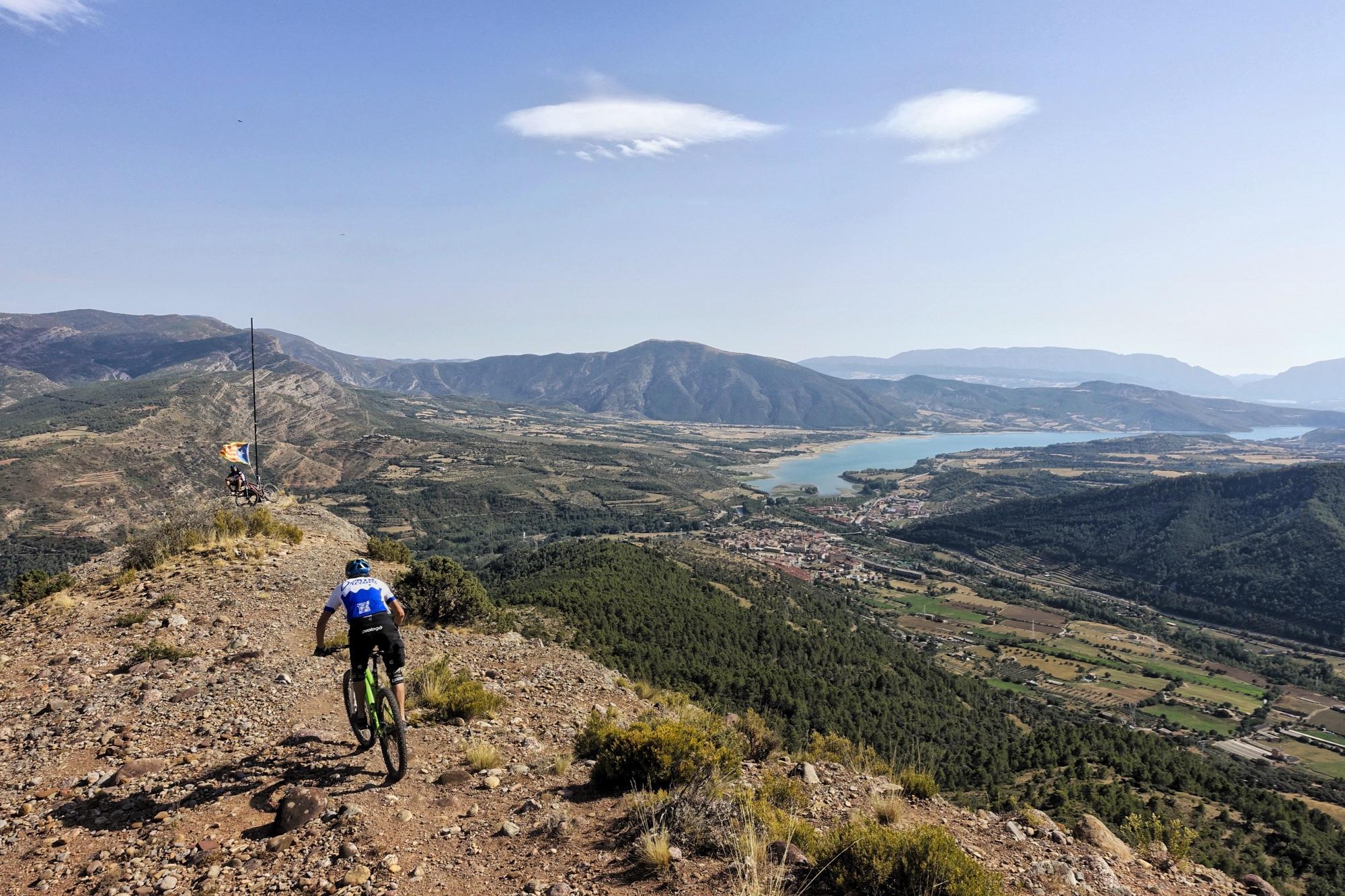 A mountain biker descends a rocky trail on a ridgeline, surrounded by a scenic landscape featuring rolling hills, a river, and a small town below. The sky is clear with a few wispy clouds, and the area is lush with green trees and vegetation. Cresta De Gelat mountain bike trail.