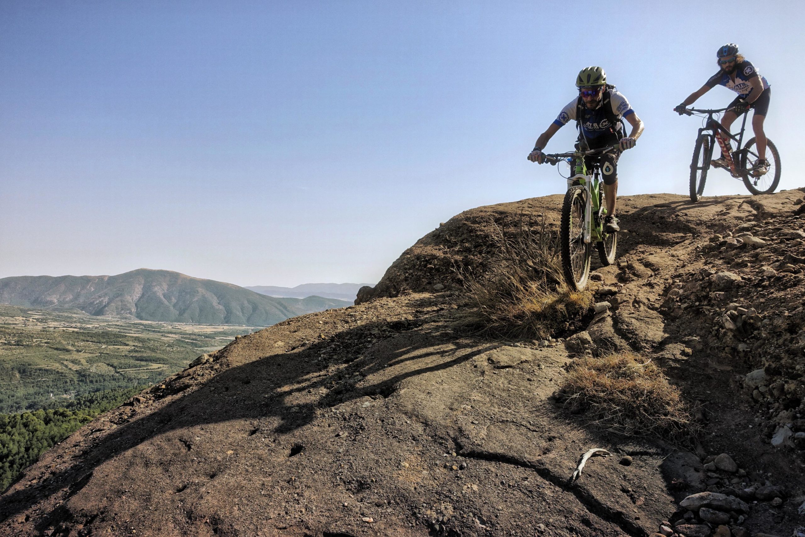Two mountain bikers navigate a rocky terrain at the edge of a cliff, with a scenic view of rolling hills and valleys in the background under a clear blue sky. Cresta De Gelat mountain bike trail.