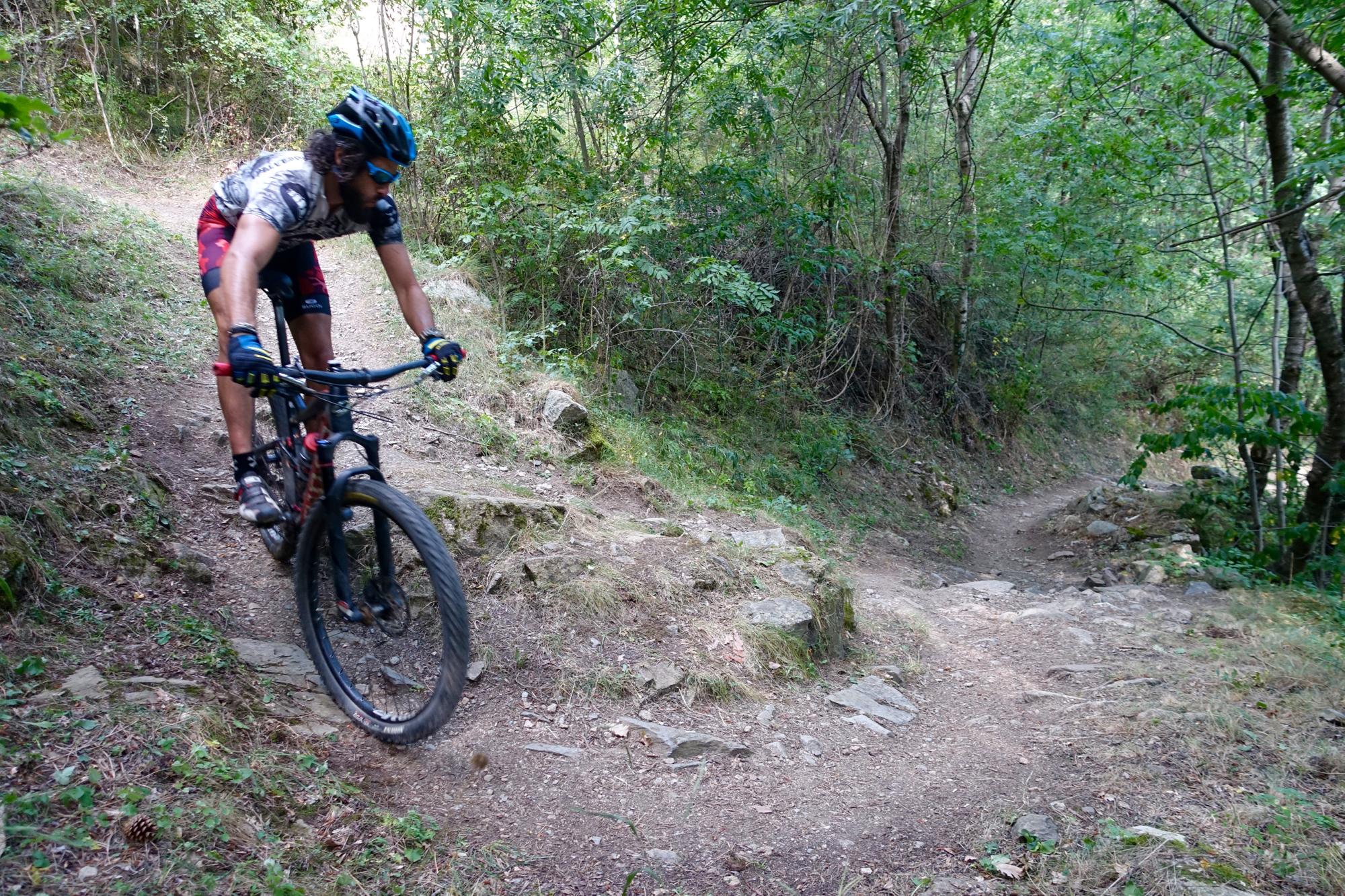 A mountain biker navigating a rocky trail surrounded by lush greenery. The cyclist is wearing a helmet, sunglasses, and athletic gear, leaning forward on the bike as they maneuver through a bend in the path. The terrain features dirt, stones, and patches of grass, highlighting an adventurous outdoor setting. Fontpedrouse mountain bike trail.