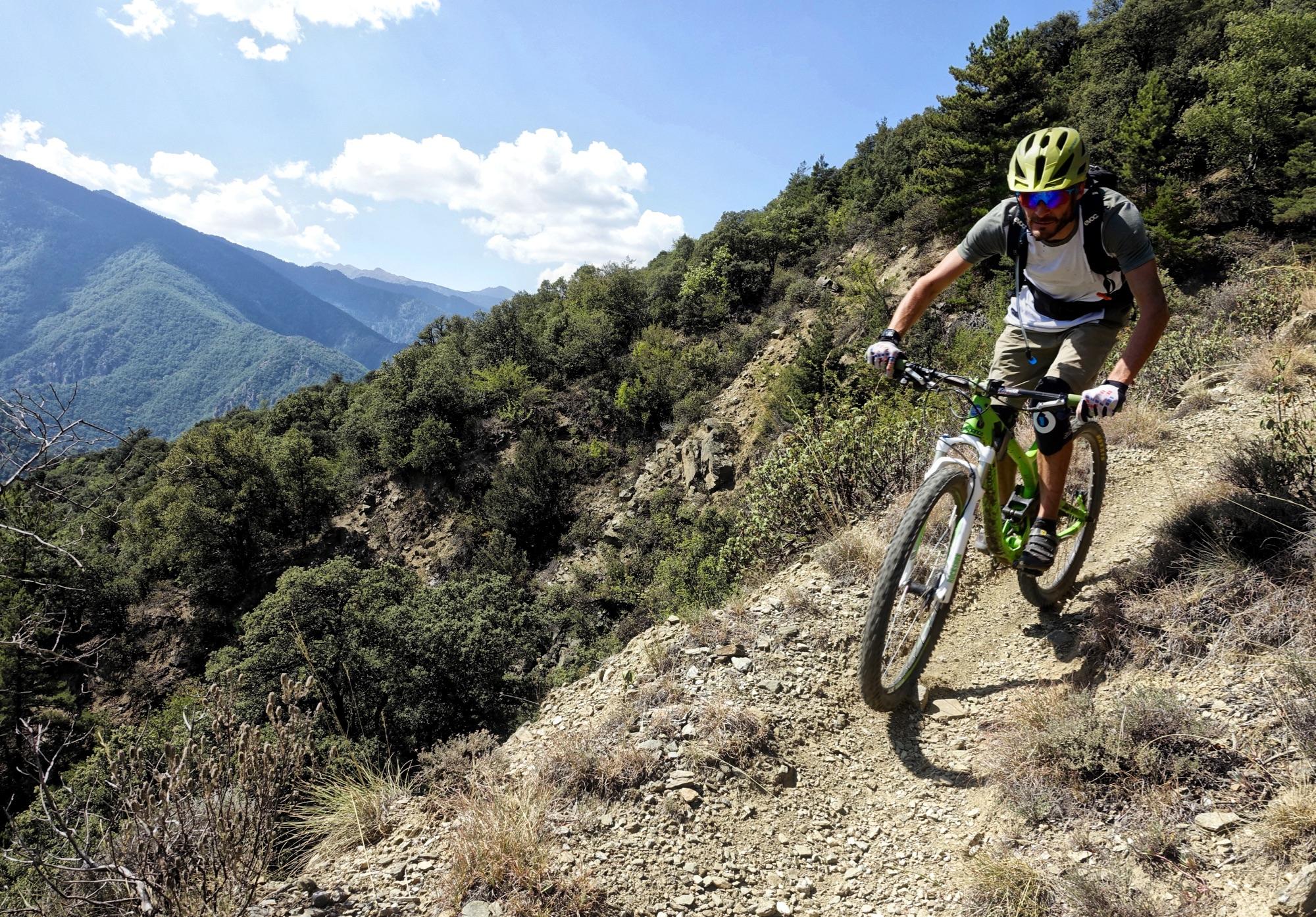 A mountain biker navigating a rocky trail surrounded by lush greenery and mountains under a bright blue sky with scattered clouds. Les Balcons du Tet mountain bike trail.