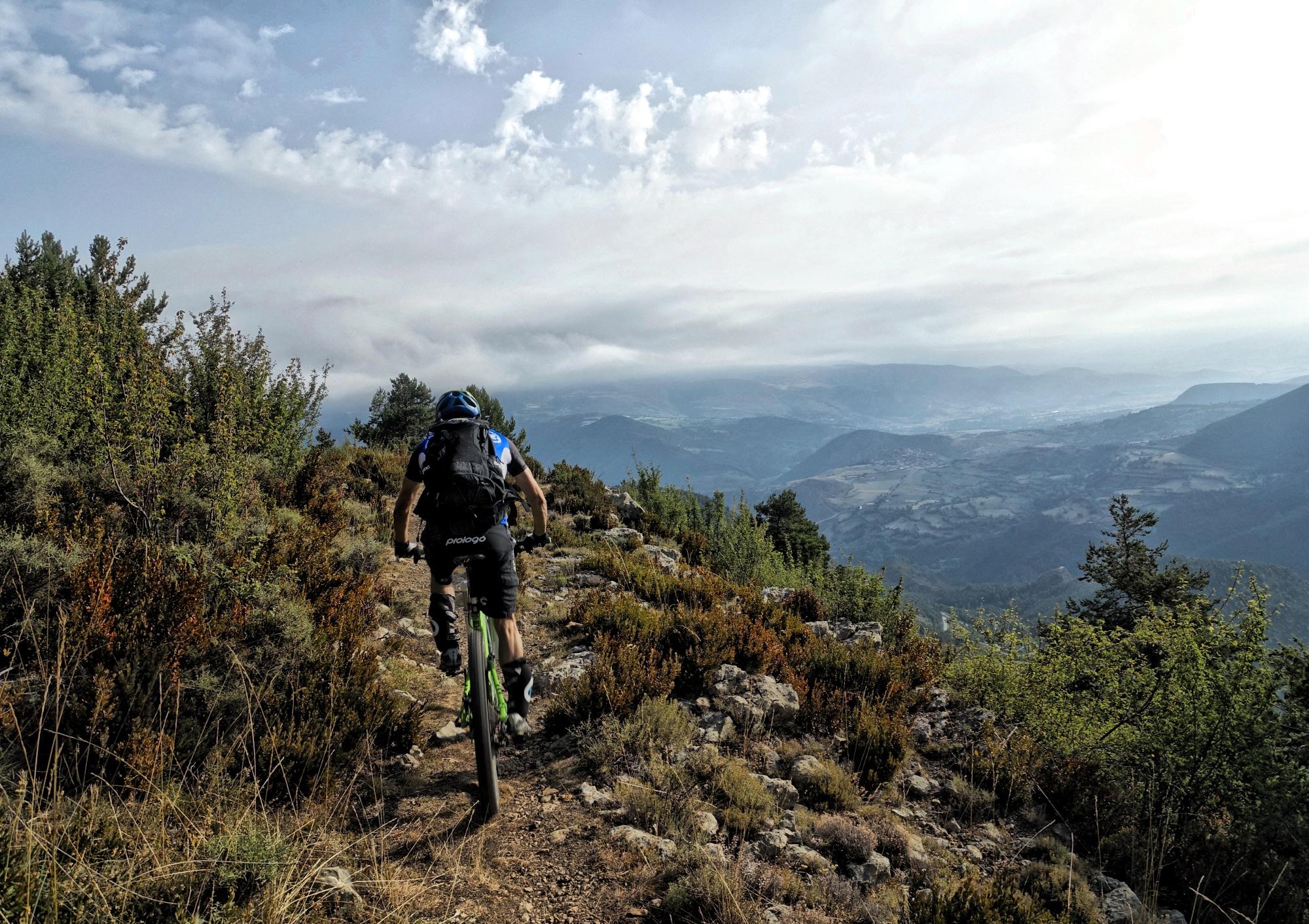 A mountain biker riding along a rugged trail surrounded by lush greenery and rocky terrain, with a stunning mountainous landscape visible in the background under a partly cloudy sky. Els Miradors D