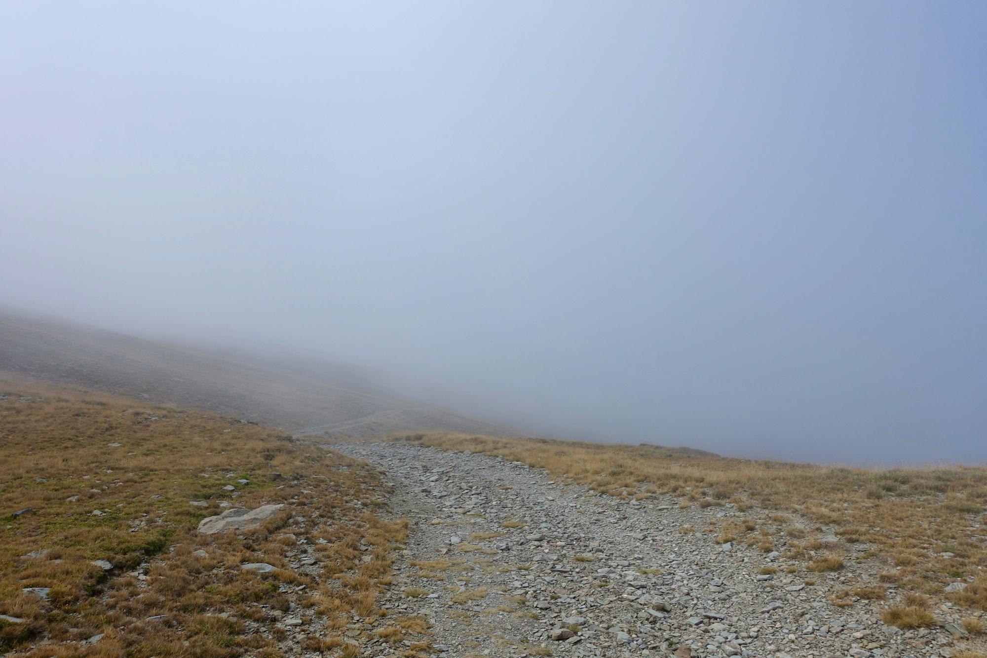 A misty landscape with a gravel path winding through gentle hills covered in dry grass and stones, partially obscured by fog. The atmosphere appears serene and quiet, evoking a sense of solitude and mystery. The Thief's Path / Pas dels Llaeres mountain bike trail.