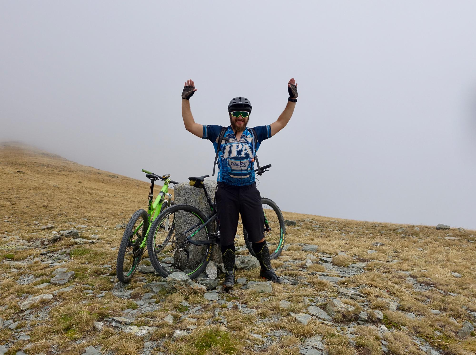 A mountain biker celebrating with arms raised, standing next to two bicycles on a rocky, grassy terrain. The background is a foggy landscape, suggesting a high-altitude environment. The biker is wearing a blue jersey, gloves, and a helmet, and appears to be enjoying the outdoor adventure. The Thief's Path / Pas dels Llaeres mountain bike trail.