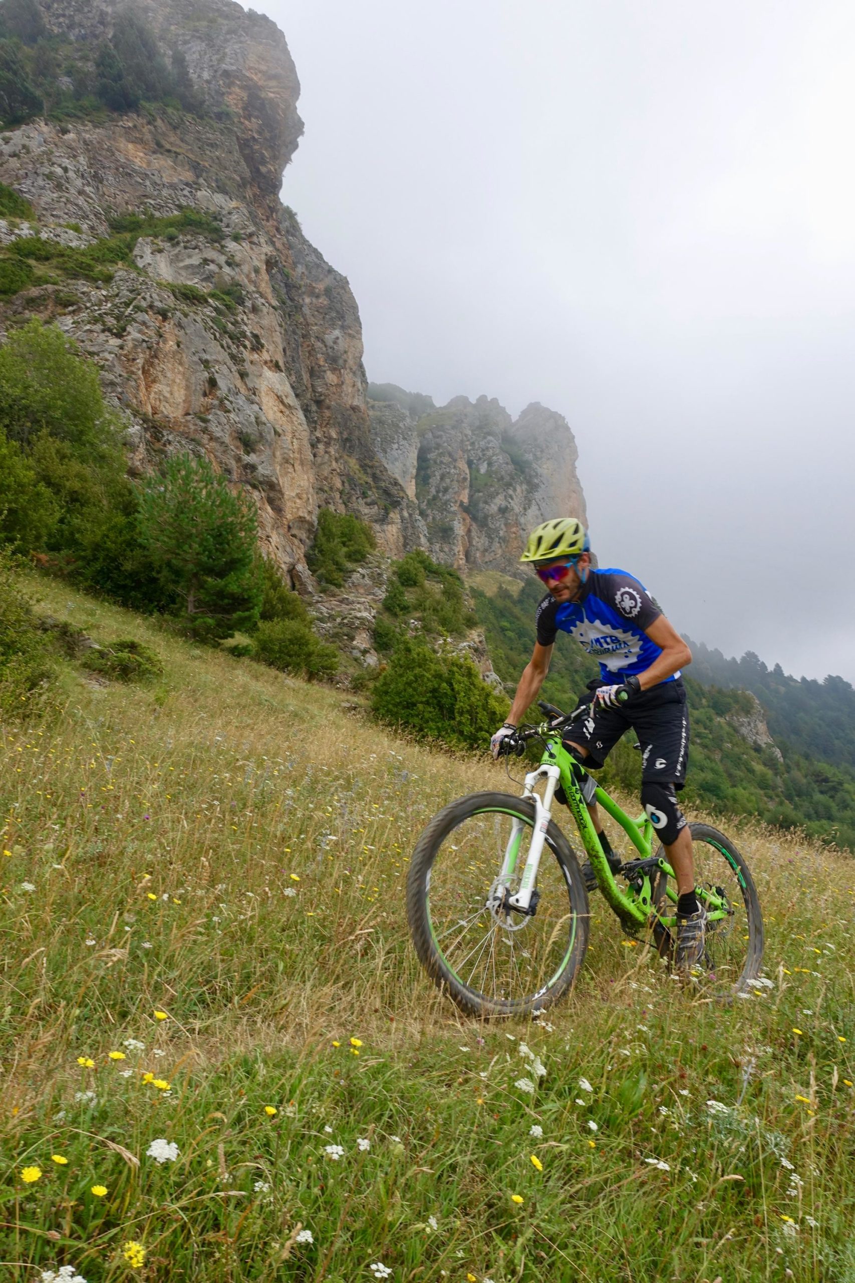 A mountain biker in a blue jersey and yellow helmet navigates a grassy hillside with wildflowers, surrounded by rocky cliffs on a foggy day. The Thief