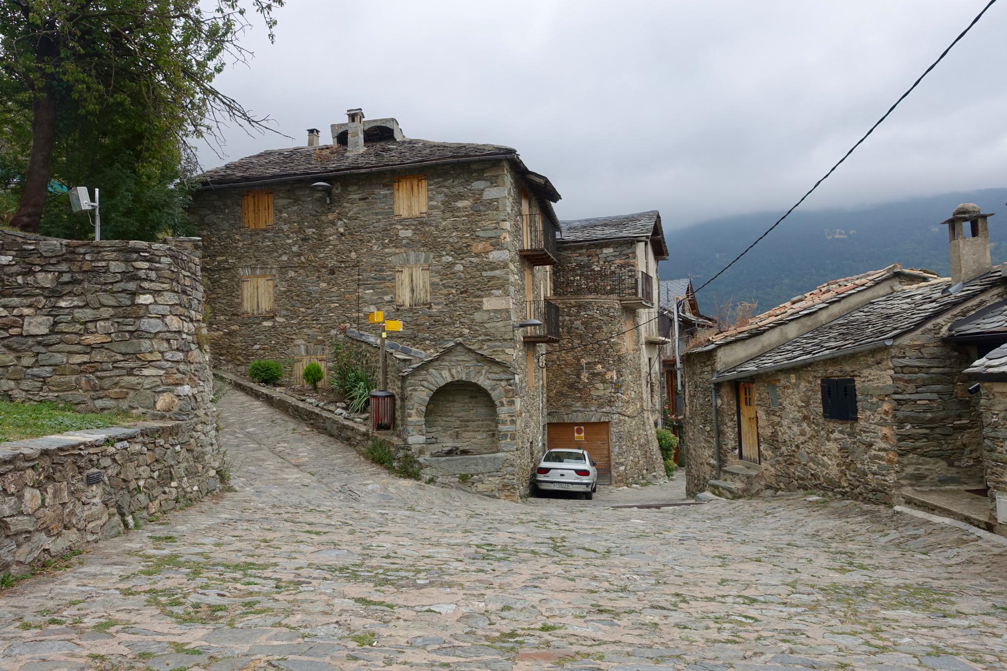 A cobblestone street winding through a picturesque village featuring rustic stone buildings with wooden shutters. A parked white car is visible at the curve of the road, and greenery can be seen along the pathway. The scene is set against a backdrop of cloudy skies and mountains in the distance. The Thief's Path / Pas dels Llaeres mountain bike trail.