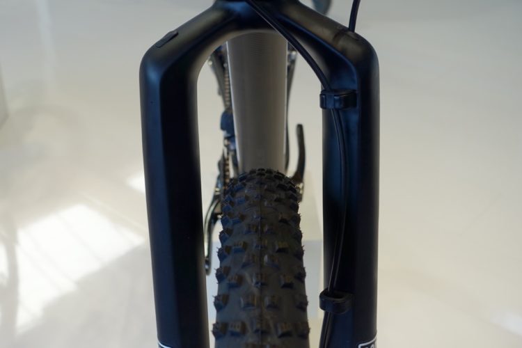 Close-up view of a mountain bike's front fork and tire, highlighting the sturdy frame and tread pattern of the tire. The background is a smooth, light-colored surface.