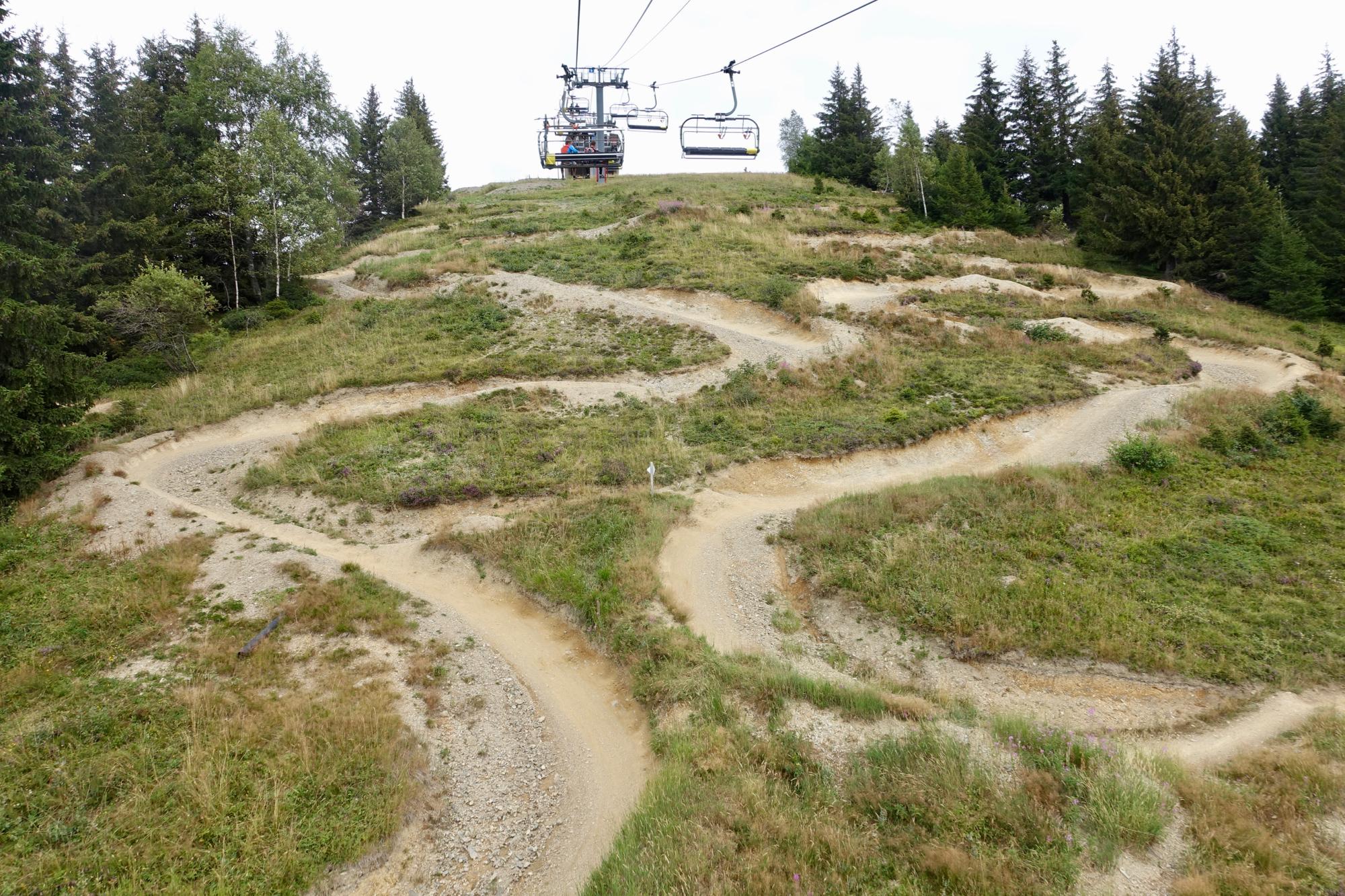 A scenic view of a winding mountain bike trail meandering down a grassy hill, with a chairlift visible in the background surrounded by lush green trees. The trail features a mix of dirt and gravel, showcasing the natural terrain. Les Gets Bike Park mountain bike trail.